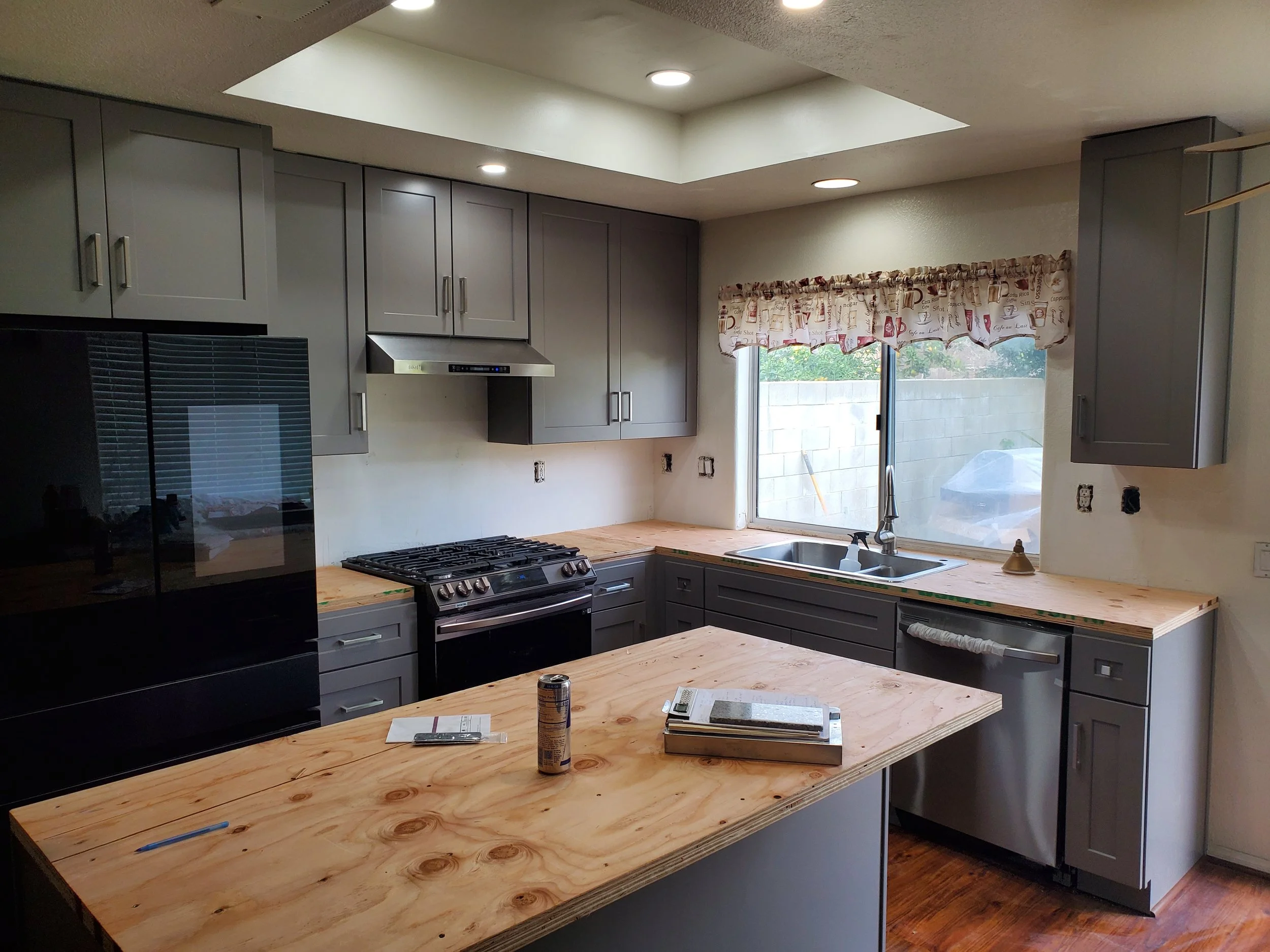 Kitchen with grey cabinets, a wooden countertop, a stove, a black refrigerator, a window with a coffee-themed curtain, and a skylight ceiling.