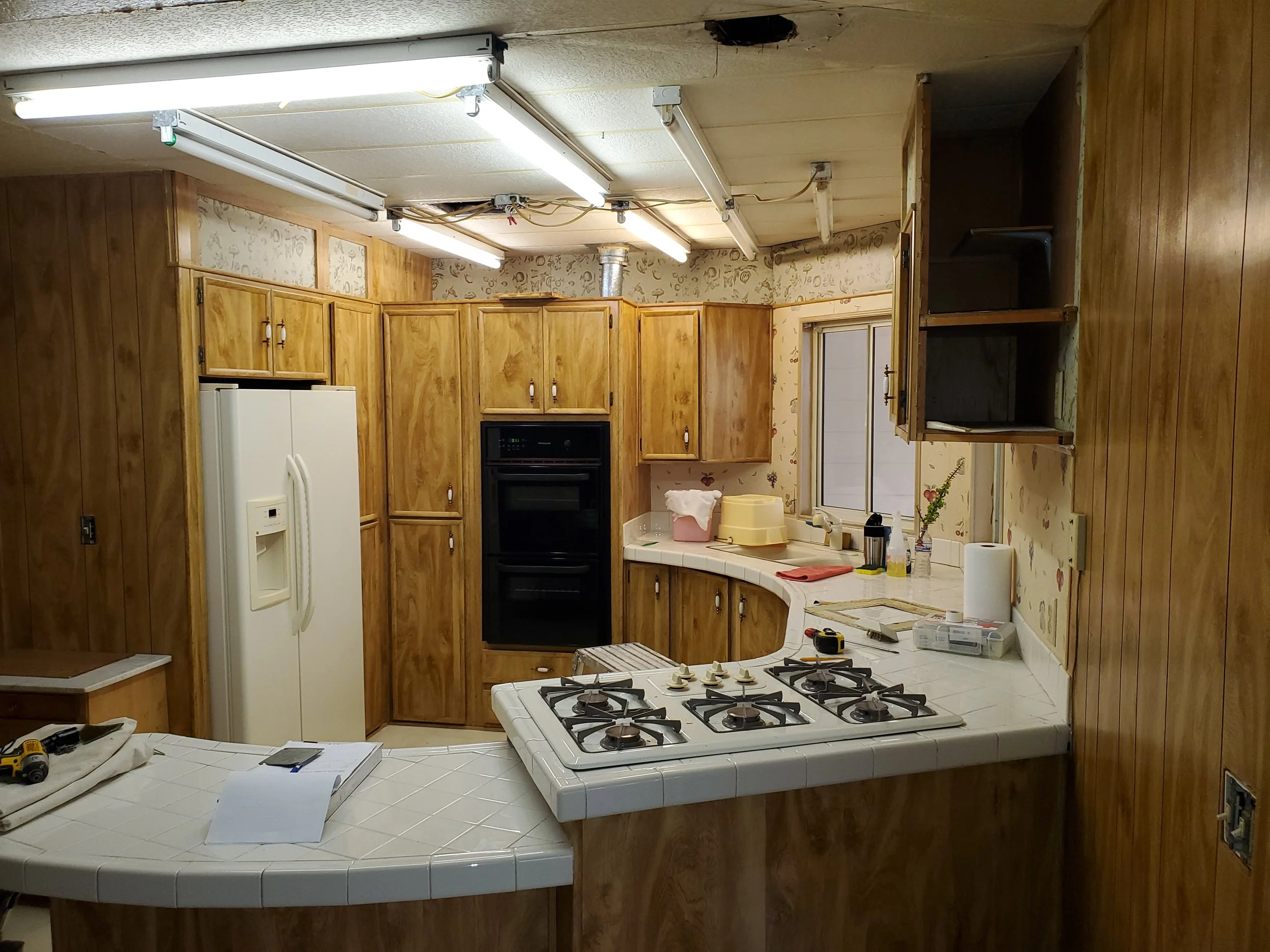 Kitchen with wooden cabinets, white appliances, a stove, a window, and a partially torn ceiling with fluorescent lights.