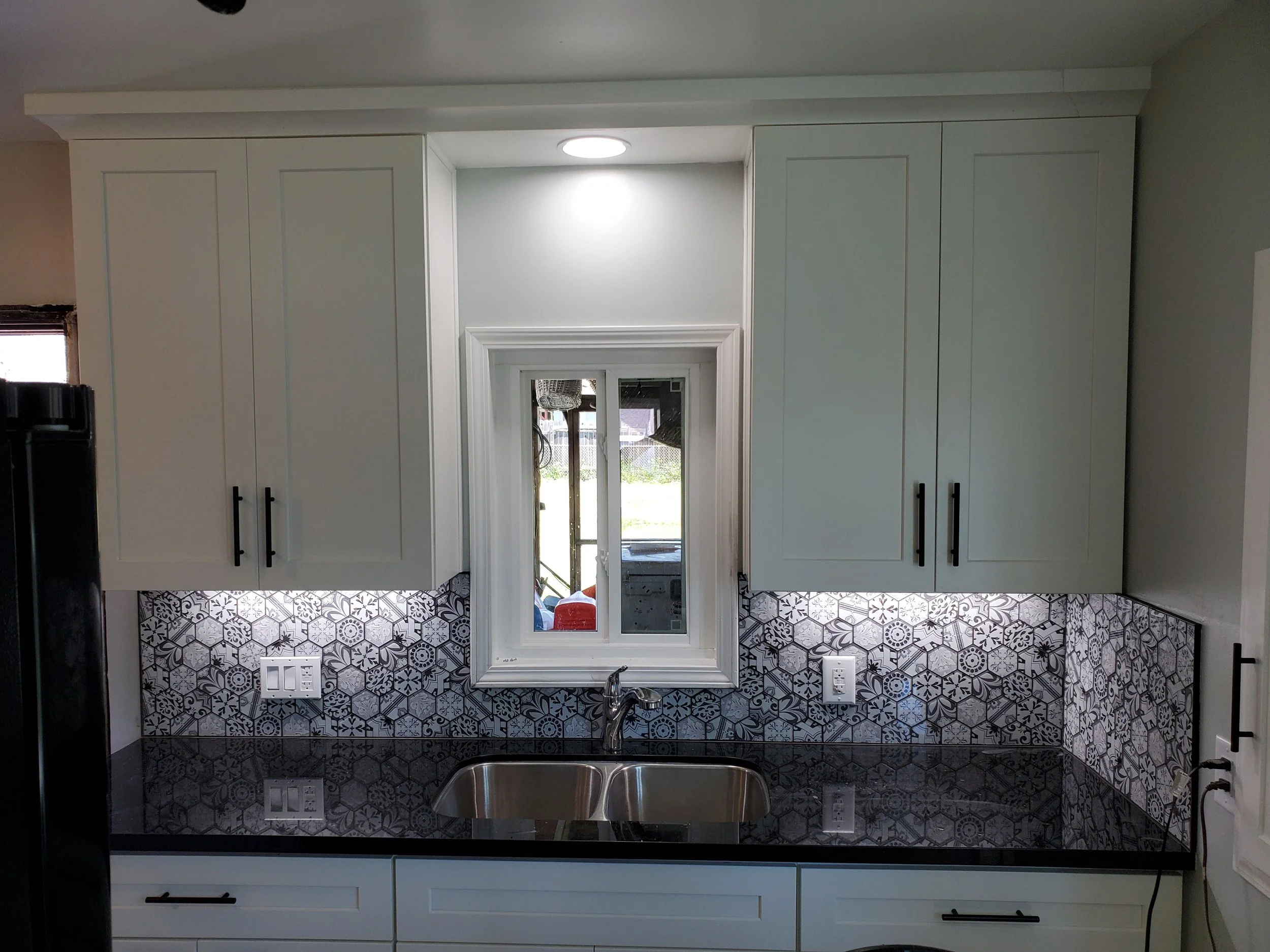 White kitchen cabinets above a window with a decorative black and white tile backsplash, a stainless steel sink, and a black granite countertop.