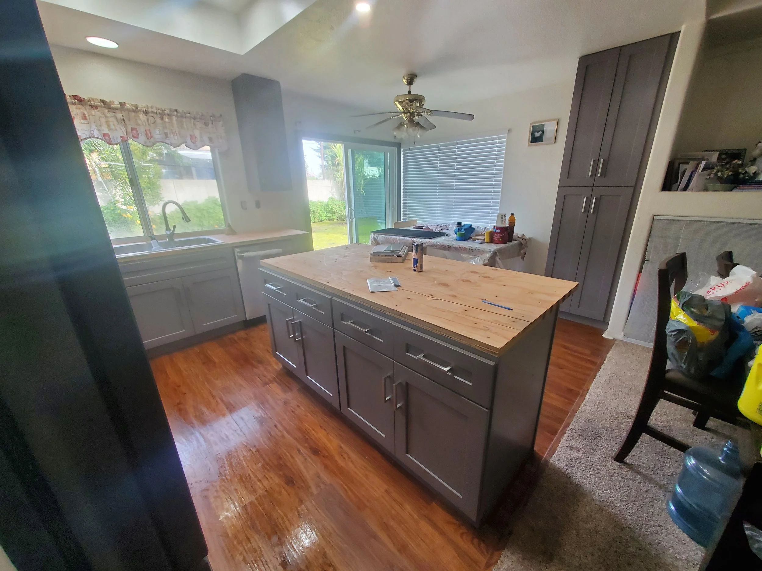 A kitchen with wooden floors, gray cabinets, a countertop island, a ceiling fan, large windows with blinds and a sliding glass door leading to a backyard, and various items and clutter on the countertop and dining table.
