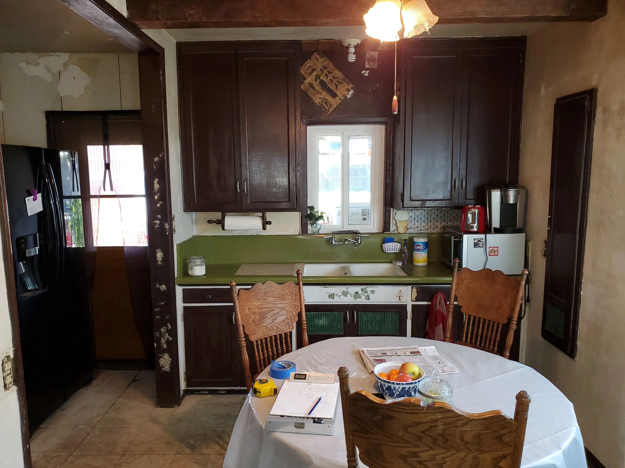 A kitchen with dark wooden cabinets above a sink and a small window. There is a table with a white tablecloth and a bowl of fruit, with chairs around it. The kitchen appears worn, with peeling paint on the wall, and various appliances on the counter.