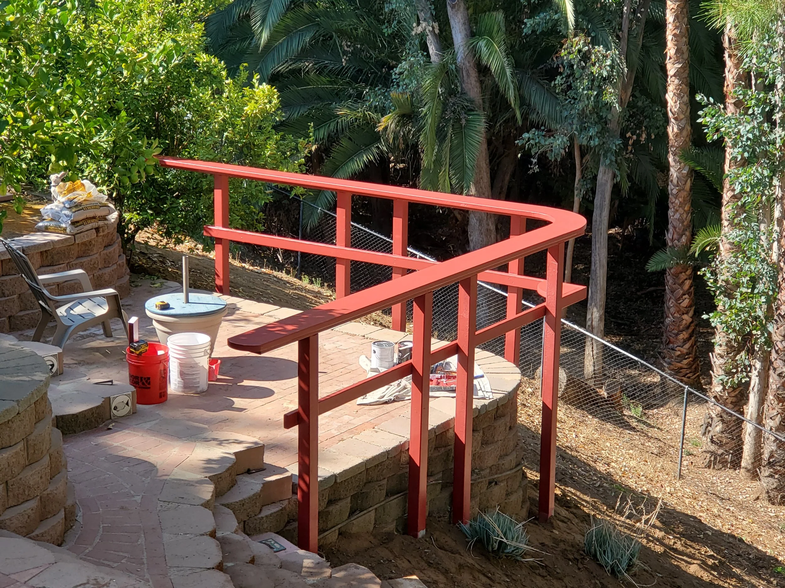 A backyard patio area with a red wooden railing, outdoor furniture, construction materials, and lush green trees and bushes in the background.