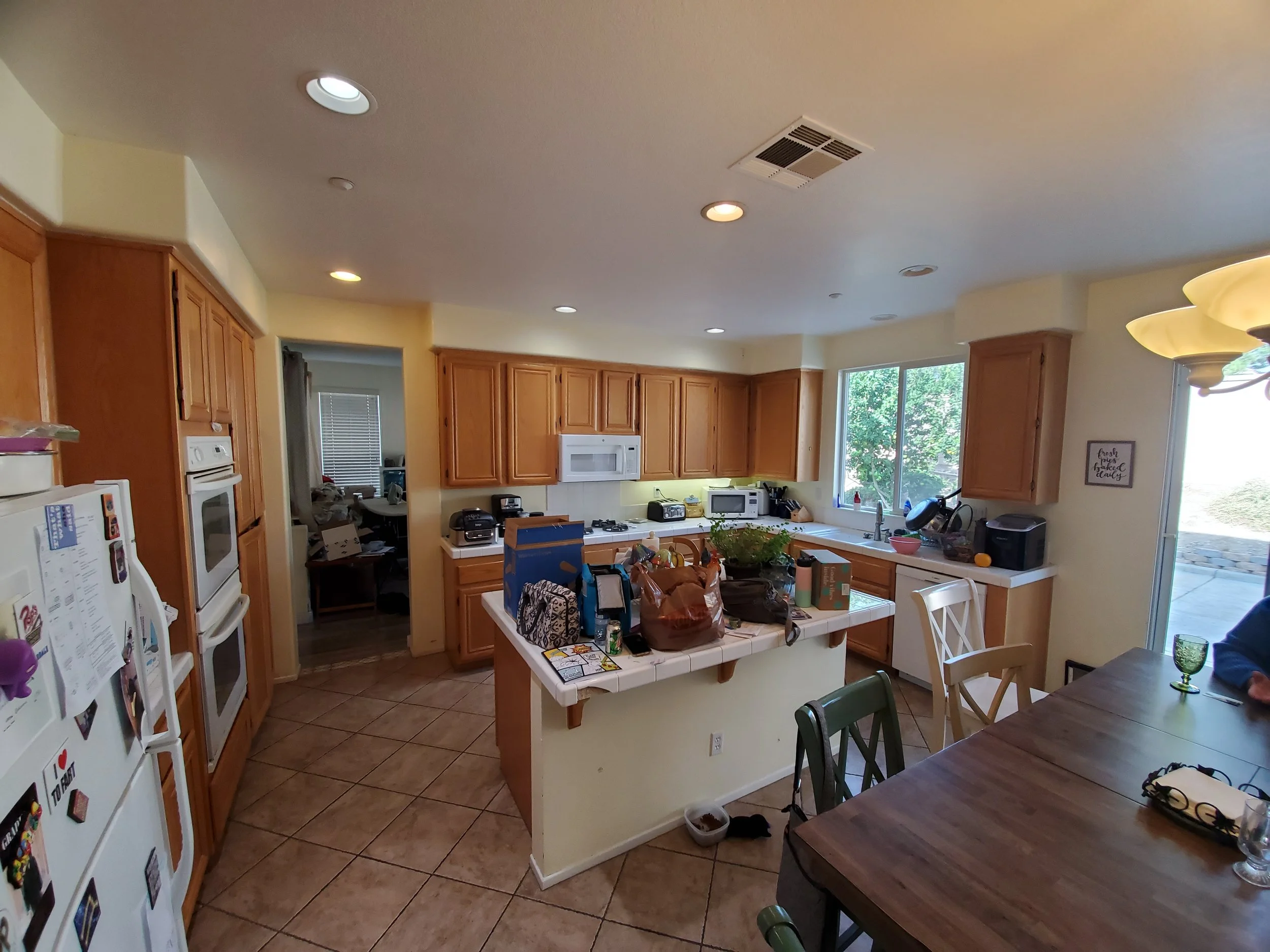 A kitchen with wooden cabinets, a white tiled countertop, a white microwave, and various appliances. There is a window with a view of trees outside, and the kitchen table has items on it, including paper, a bag, and a water glass. Recessed ceiling li
