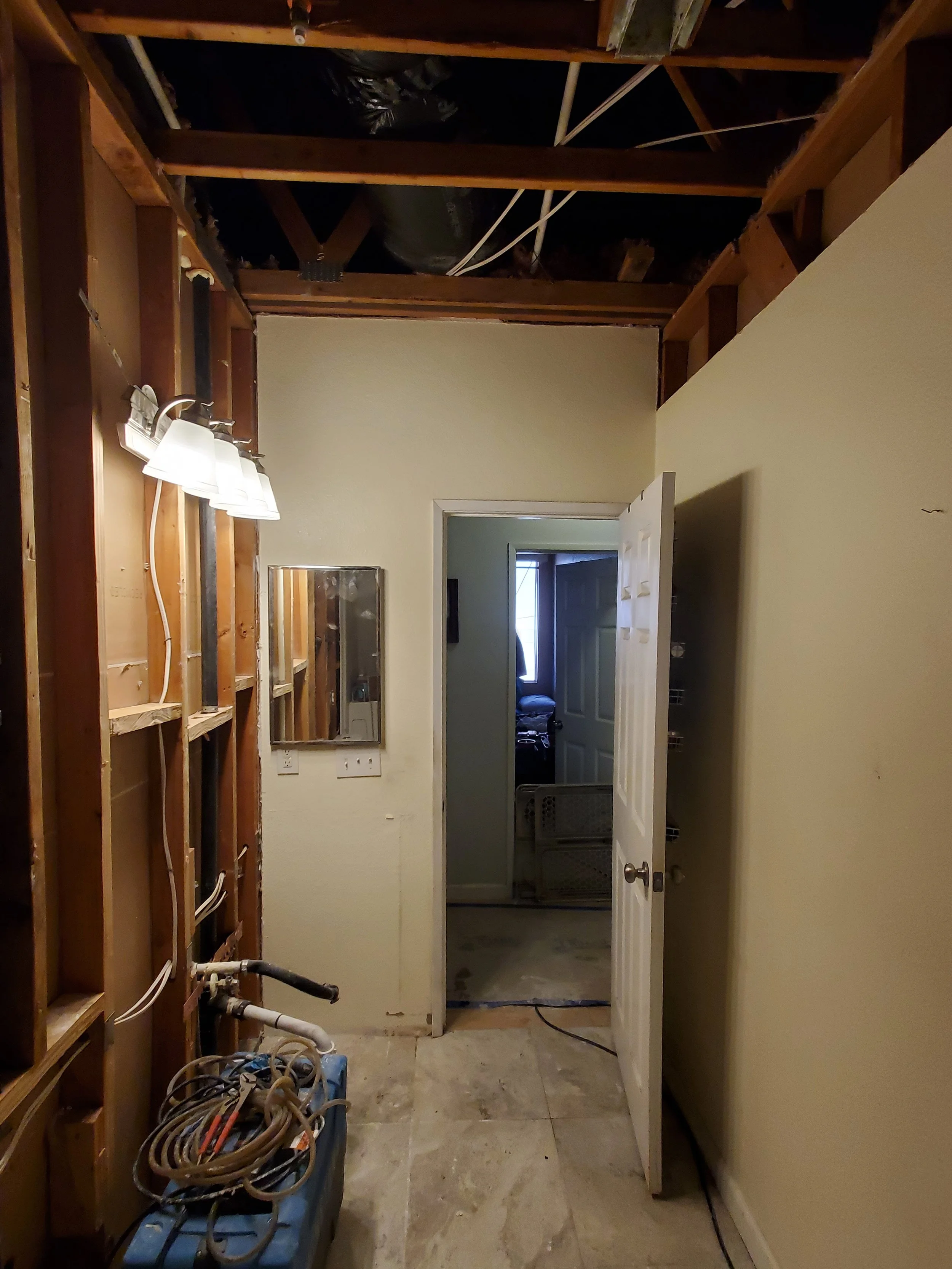 Partially unfinished room with exposed wooden framing, pipes, and electrical wires, leading into an adjacent room through a doorway, with a mirror on the wall and a box of tangled cables on the floor.