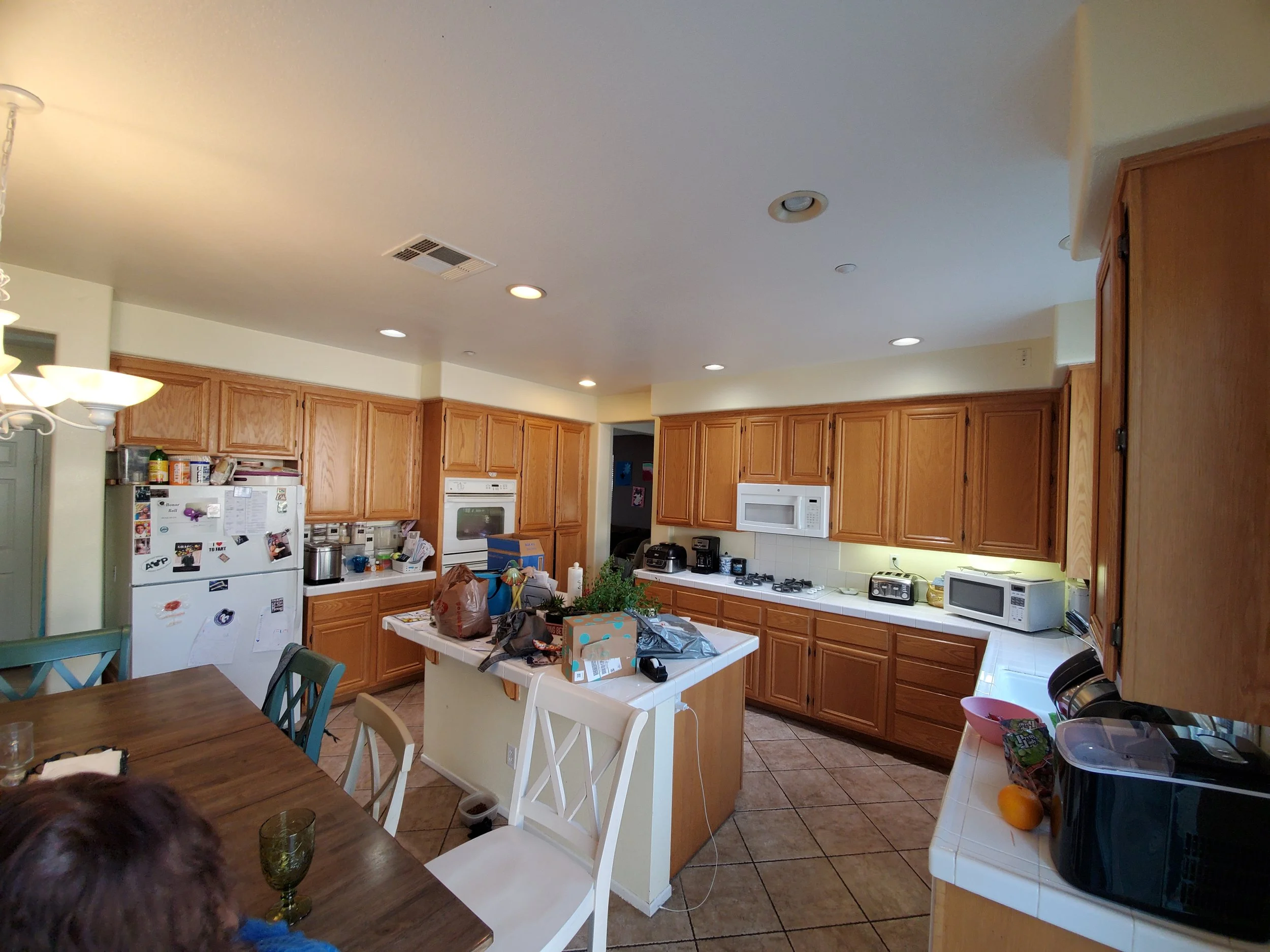 View of a busy kitchen with wooden cabinets, a white countertop, and various appliances. The kitchen table has items on it, and clutter is visible throughout the space.