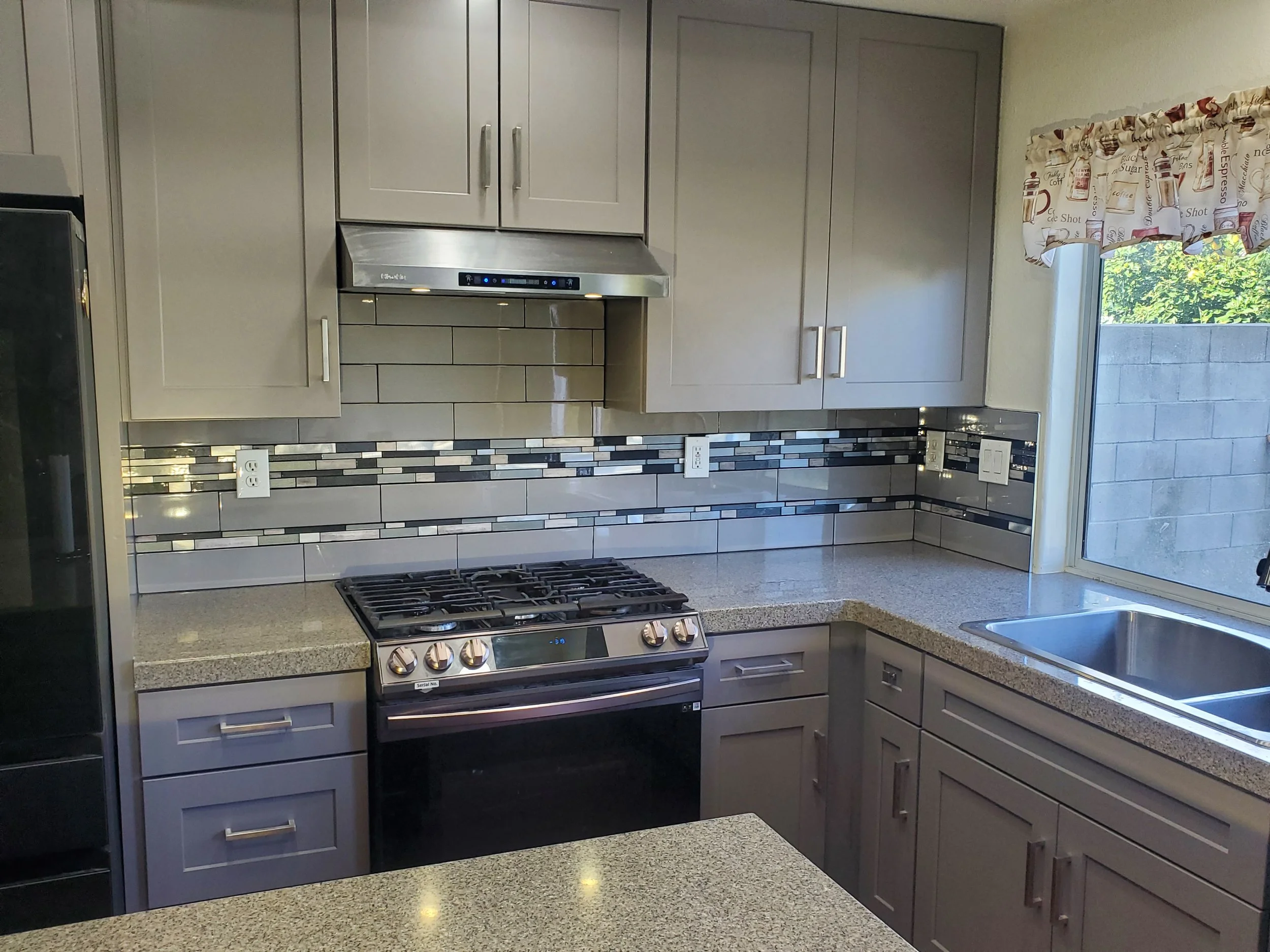 A modern kitchen with gray cabinets, a stainless steel range hood, a gas stove, a mosaic backsplash, a window with a decorative curtain, and a granite countertop.
