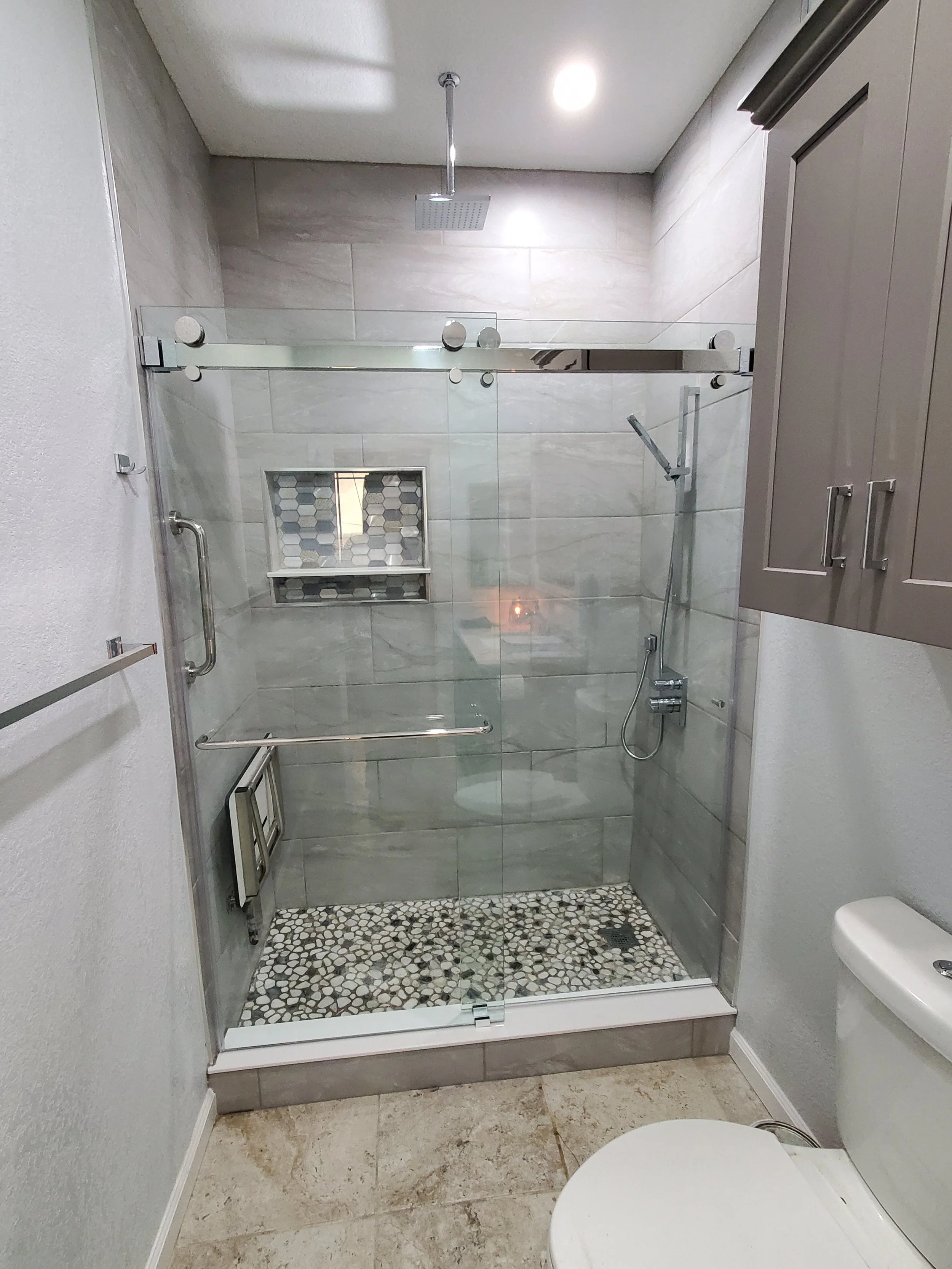 Bathroom with a walk-in shower featuring glass doors, a rainfall showerhead, and a pebble tile floor, with a beige tile wall and a built-in shower niche.