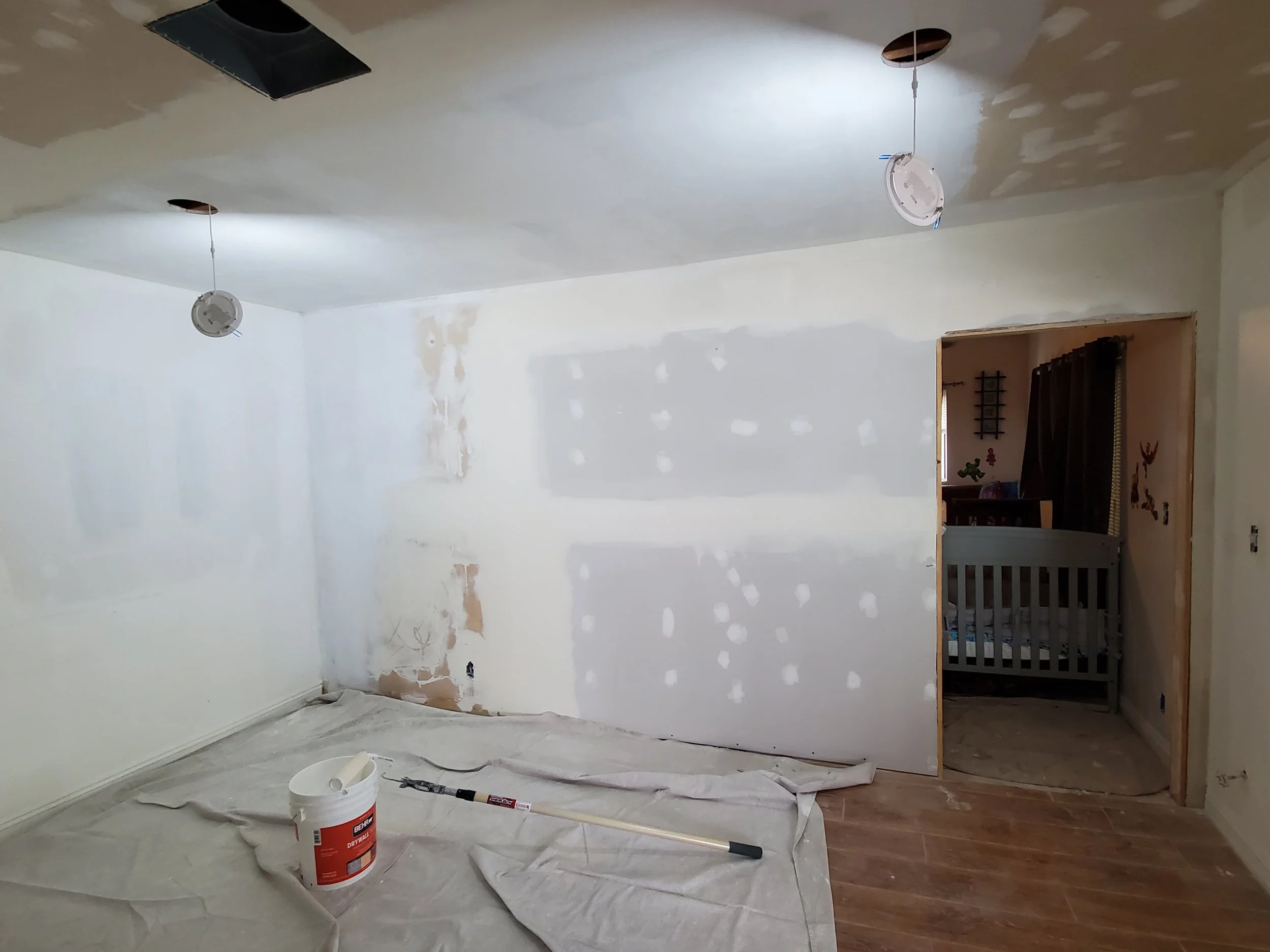 Room undergoing renovation with drywall partially painted, construction tools and supplies including a bucket, roller, and ceiling holes visible.
