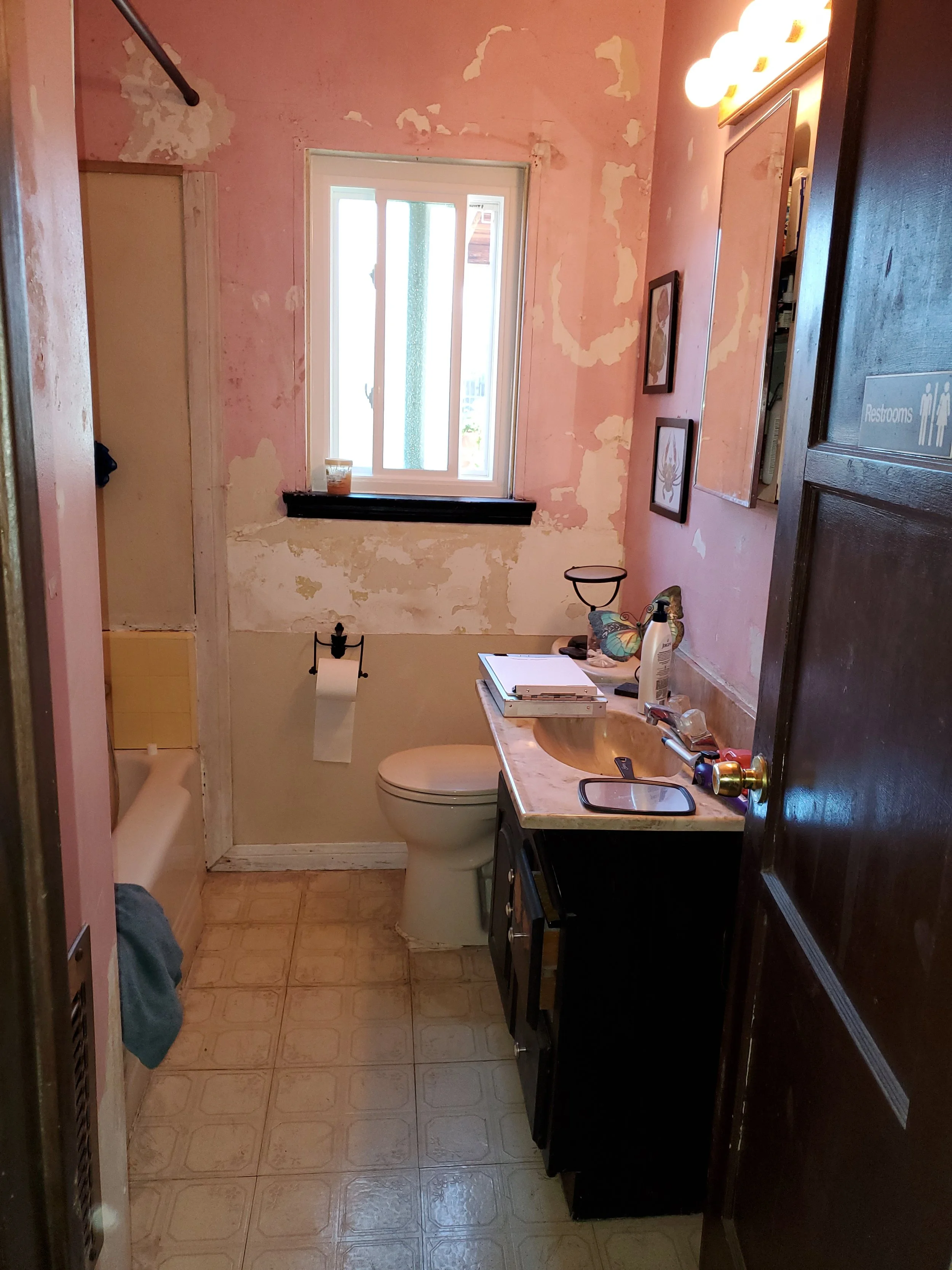 Small bathroom with peeling pink paint on walls, a window above the toilet, a black cabinet, a sink with a small countertop, a mirror with lights, framed pictures on the wall, and various toiletries on the countertop.