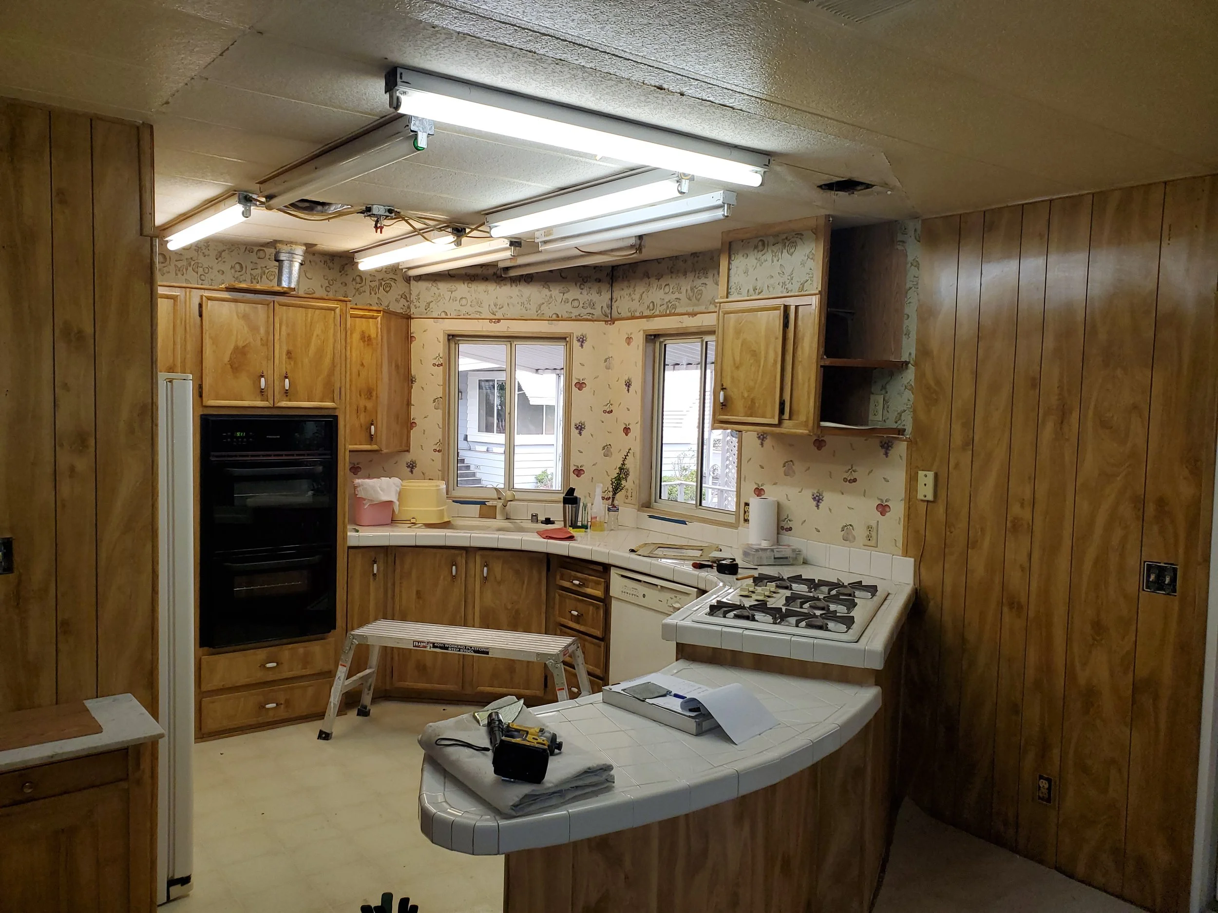 Kitchen with wood-paneled walls, fluorescent ceiling lights, and two windows. There are cabinets, a stove, a dishwasher, and a white tiled countertop. Tools and construction materials are on the countertop and floor, indicating ongoing renovation.