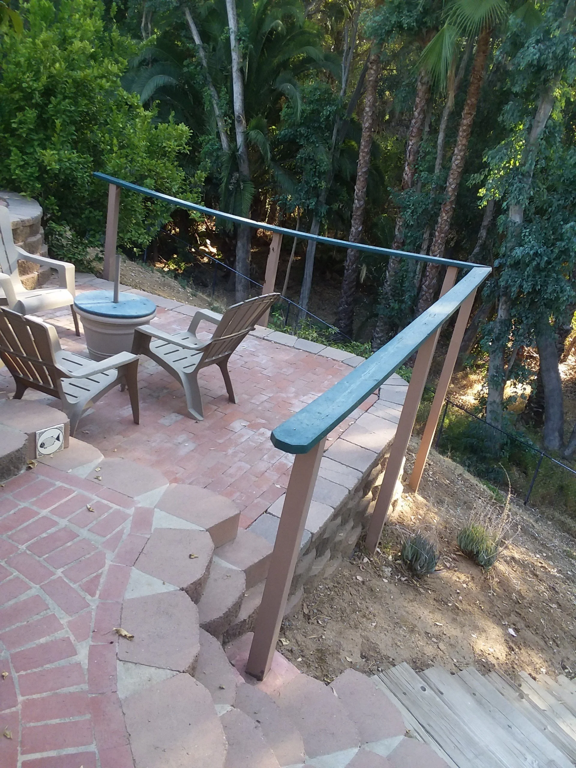 A patio area with chairs and a small table, surrounded by lush greenery and tall trees.