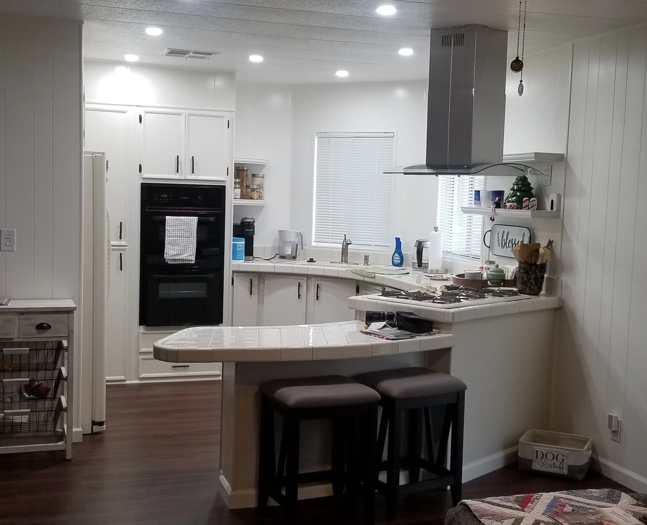 Kitchen with white cabinets, a black oven, a double sink with a window above, a small table with two black stools, decorative items on a shelf, and a white trash bin with a dog-themed design.