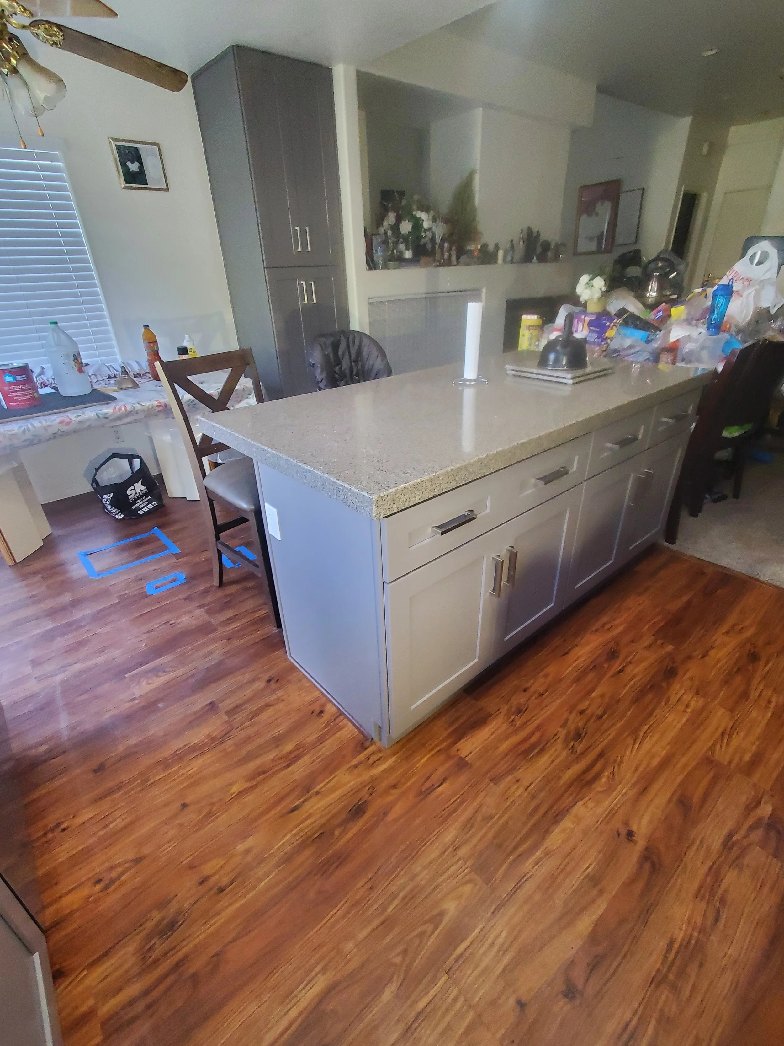 A kitchen with a beige countertop, white cabinets, and a wooden floor. There is a counter with various items on it, including a black kettle, snacks, and a plastic bag. In the background, there are flowers, a picture on the wall, and a window with bl