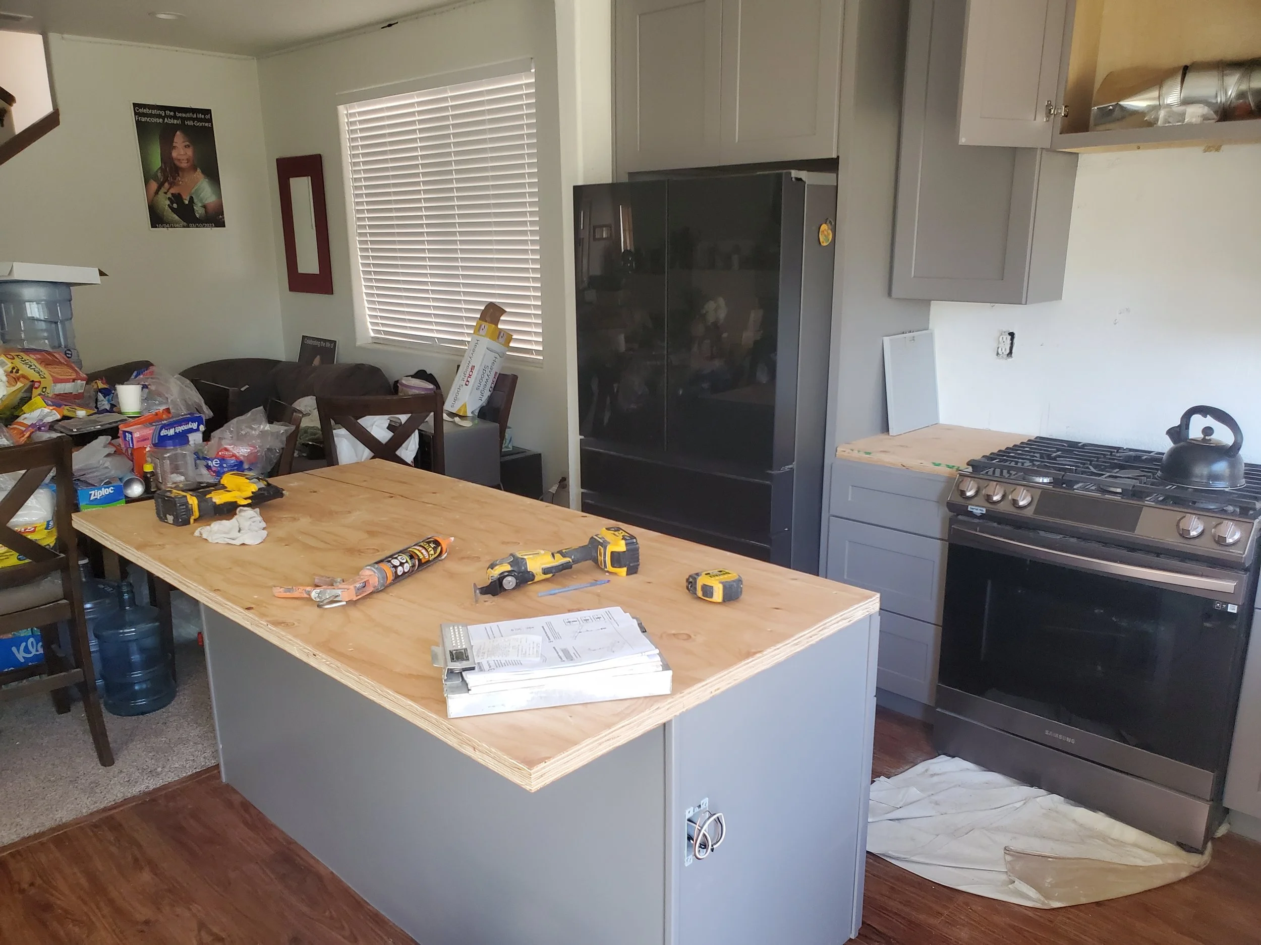 A kitchen with a light gray island counter, cluttered with tools, paper, and snacks; a black refrigerator, gray cabinets, a stove, and a window with white blinds.