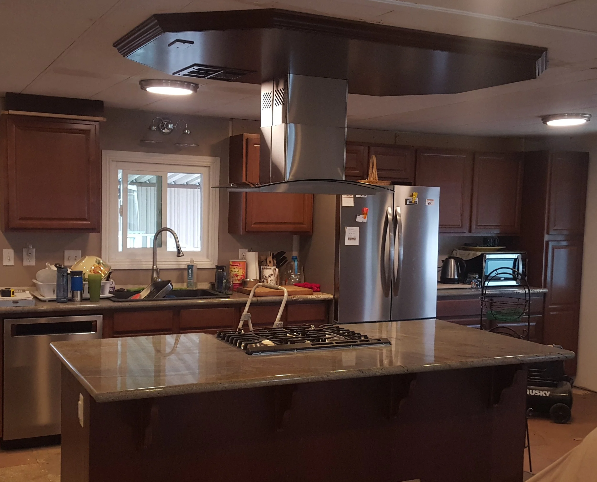 Kitchen with granite island countertop in center, stainless steel appliances, wooden cabinets, window above the sink, and various kitchen items on counters.