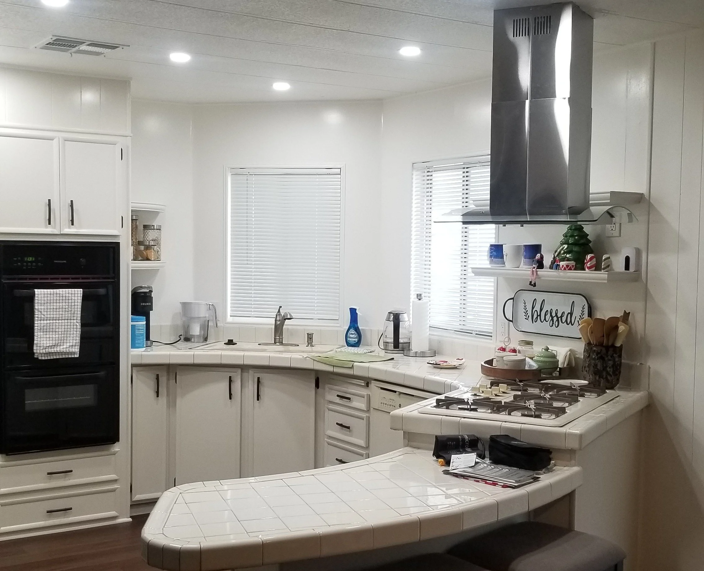 A kitchen with white cabinets, windows with blinds, a stove, and various household items on the countertops and shelves.