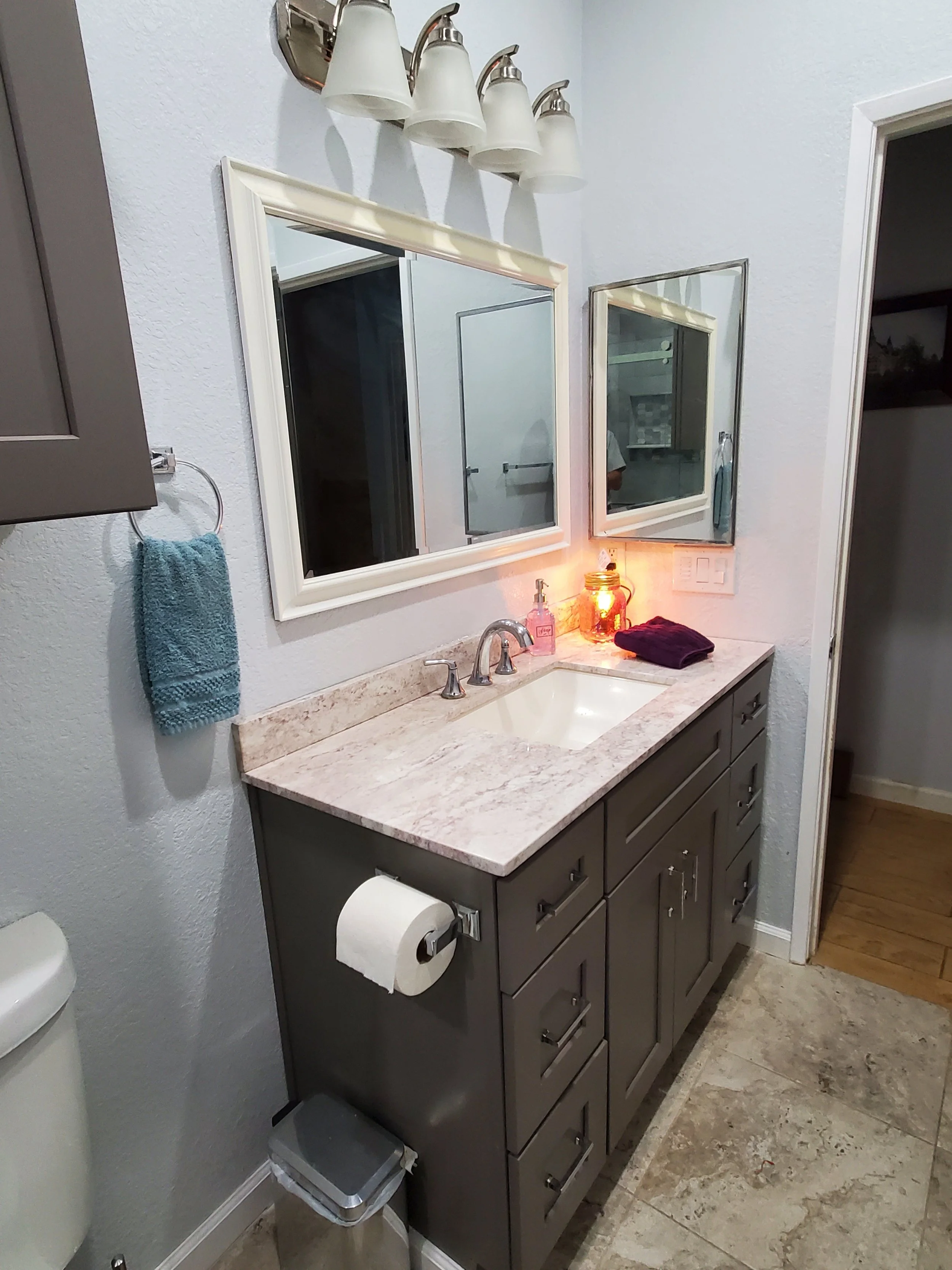 Bathroom vanity with a sink, a mirror, a wall-mounted light fixture with four lamps, a blue hand towel on a towel ring, a purple cloth, a soap dispenser, a jar with a candle inside, a paper towel holder with toilet paper, and a part of a toilet. The 