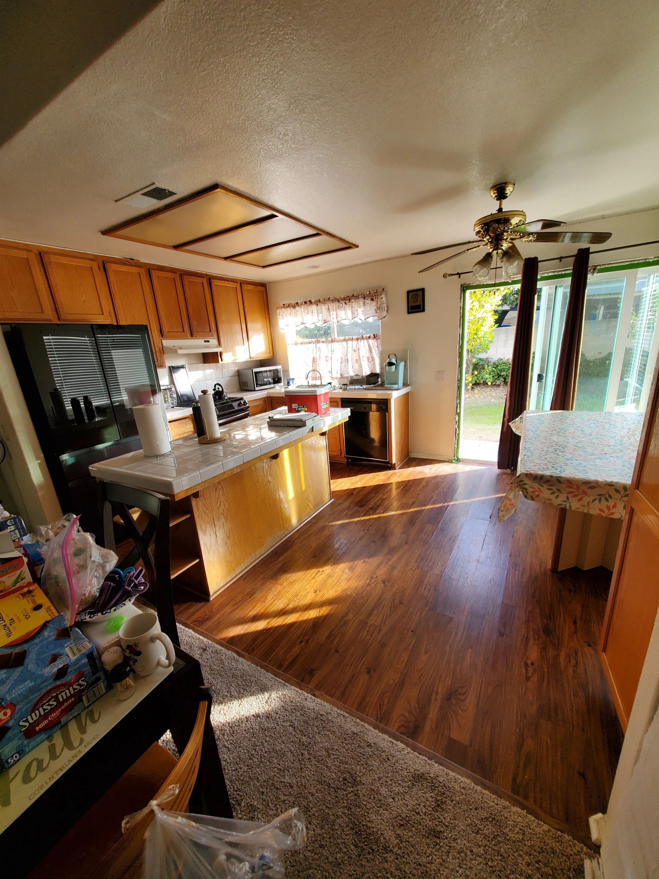 A kitchen with wooden cabinets, a ceiling fan, sliding glass door with curtains, and a dining table with a tablecloth. The kitchen has a tiled island and some kitchen appliances. There's a window with curtains and a view of a yard outside.