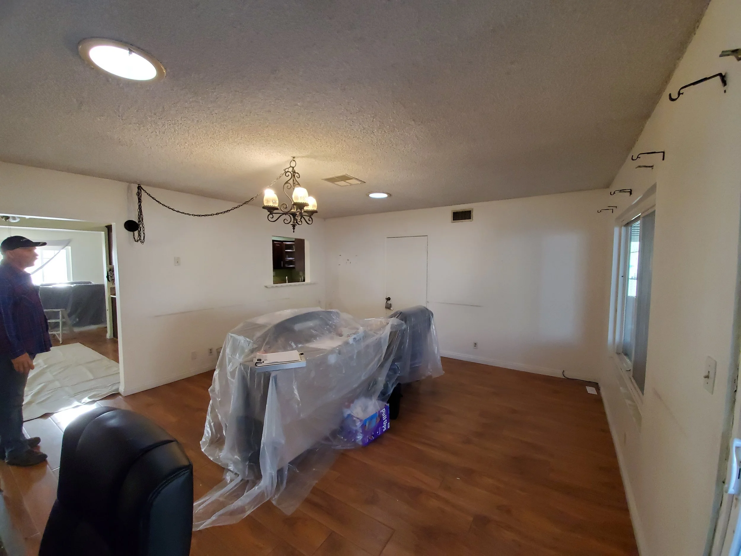 Living room with wood flooring, ceiling lights, chandelier, and hooks on the wall. Furniture covered with plastic, a person standing near the doorway, and a window with blinds.
