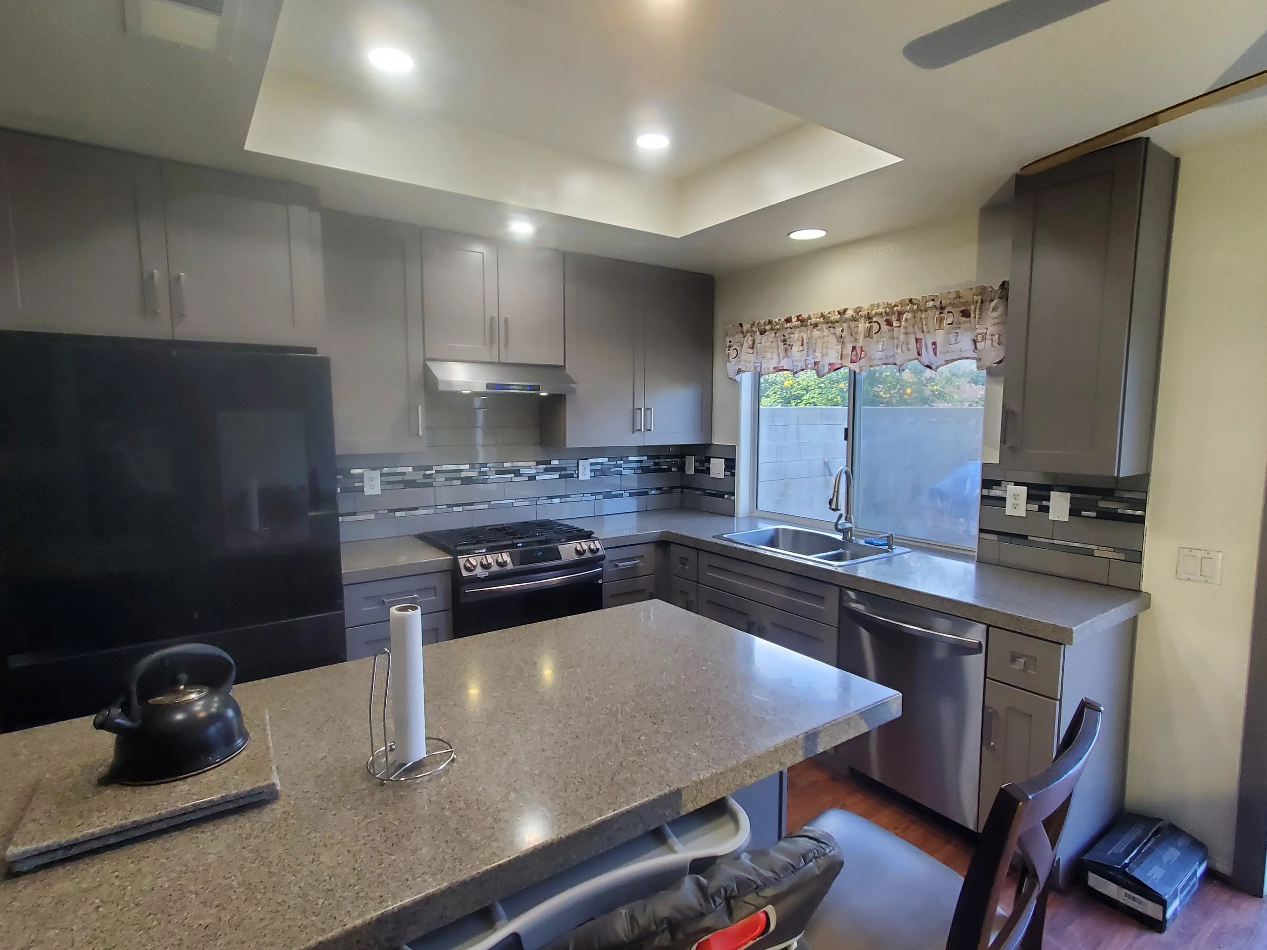 Kitchen with gray cabinets, a window with a colorful curtain, a sink under the window, a gas stove, tiled backsplash, and a countertop with a paper towel holder and a kettle.