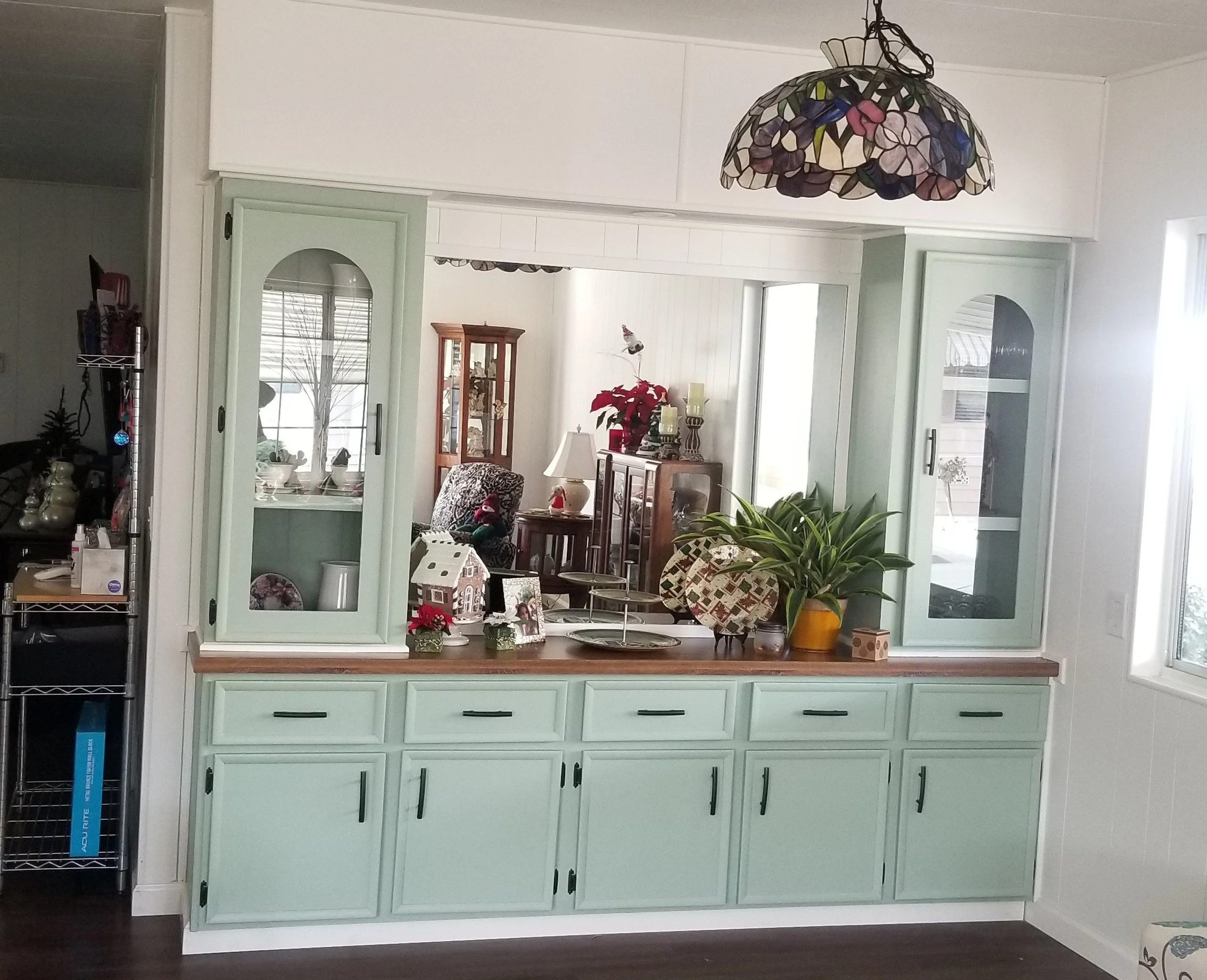 View of a light green kitchen cabinet with black handles, with various decorative items including small Christmas-themed decorations, a house-shaped ornament, and a potted plant in a yellow pot on top. The background shows parts of other rooms with f