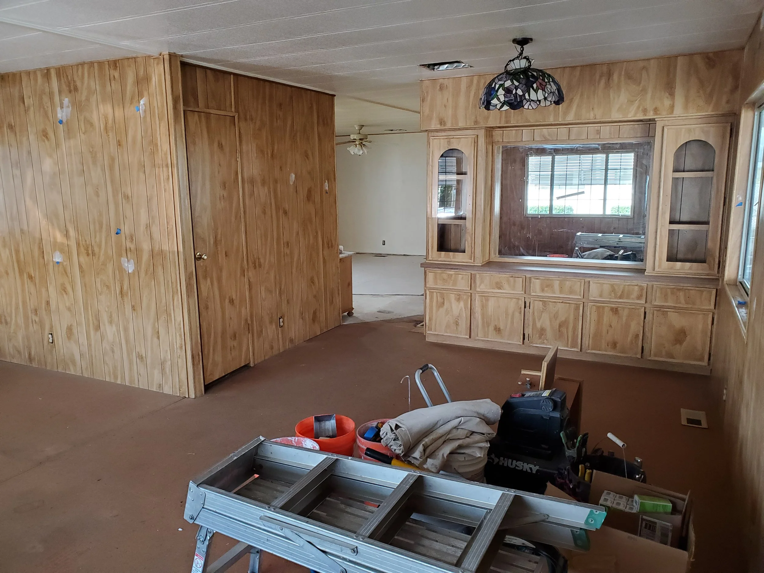 Room under renovation with wood-paneled walls, construction tools, and materials scattered on the floor, including a partially assembled metal frame, buckets, and a toolbox.