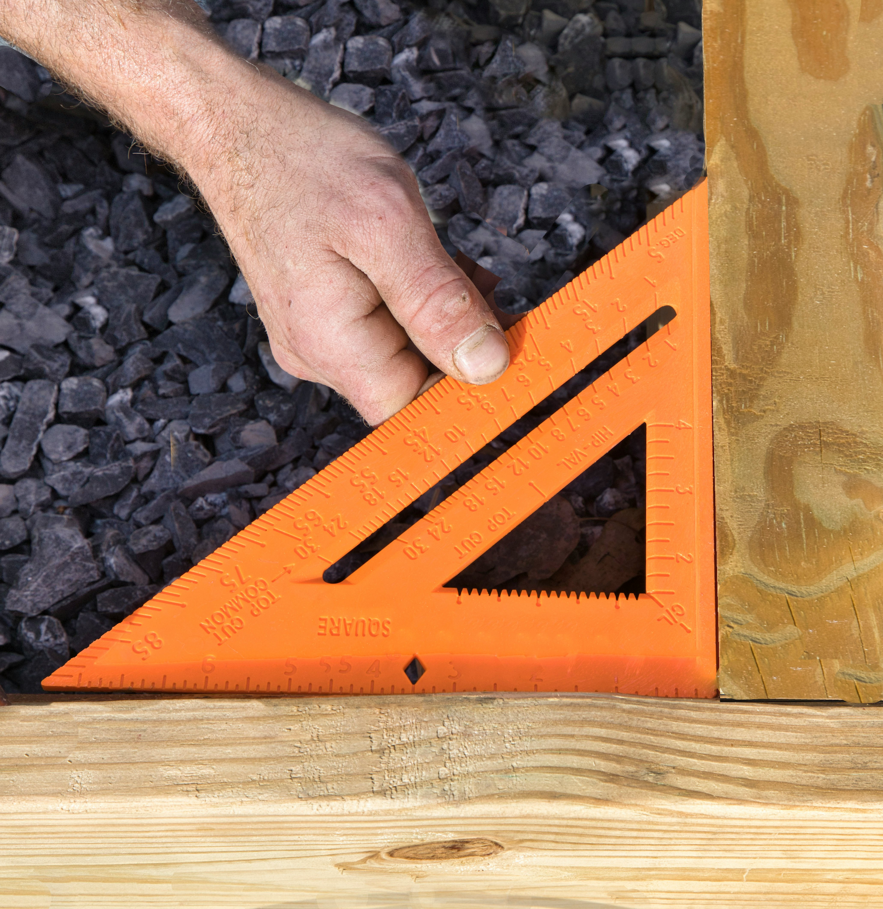 Close-up of a human hand holding an orange framing square on wooden and gravel surfaces.