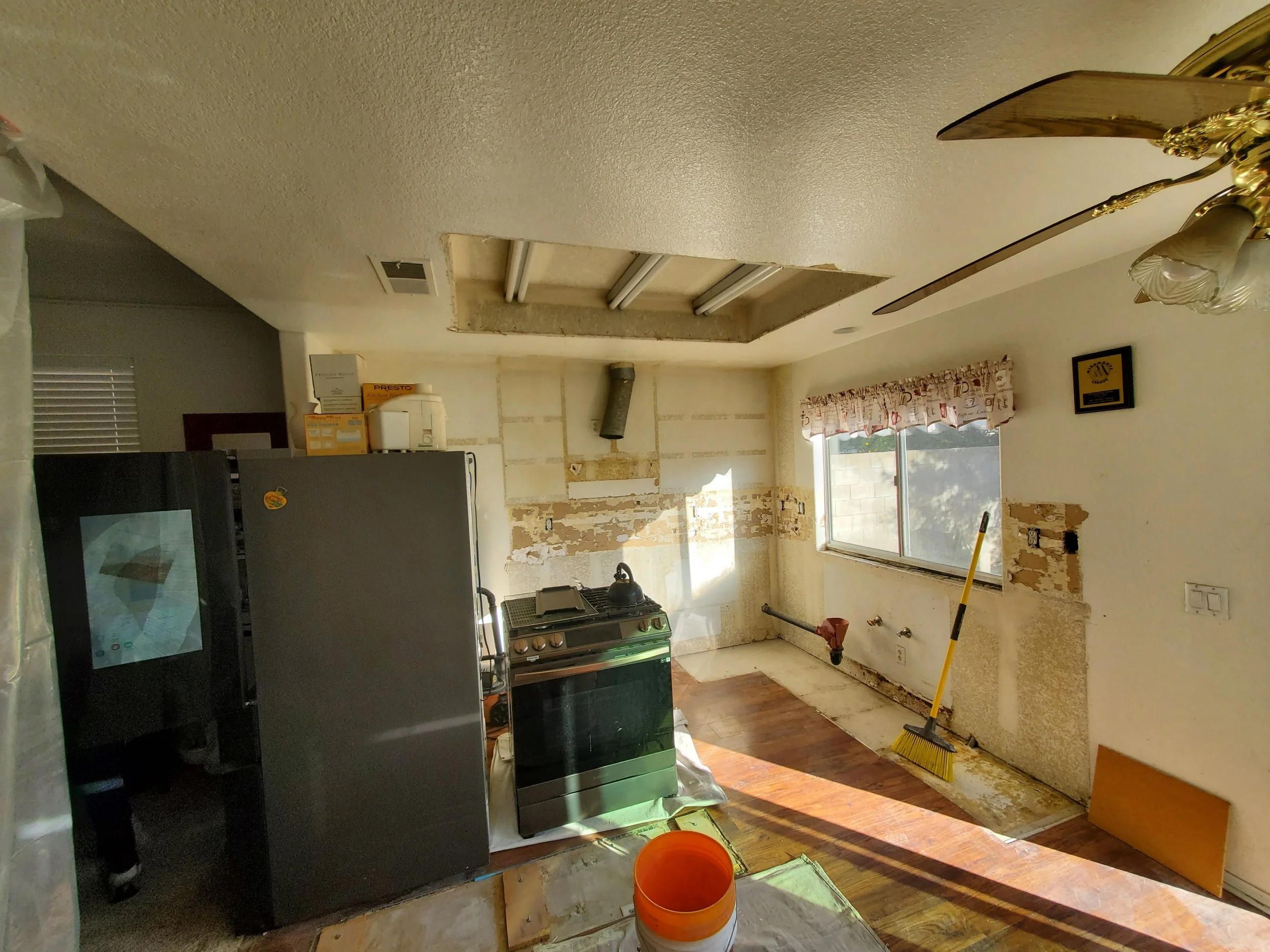 Room undergoing renovation with exposed drywall, a window with curtains, a stove, a fridge, cleaning tools, and construction materials.