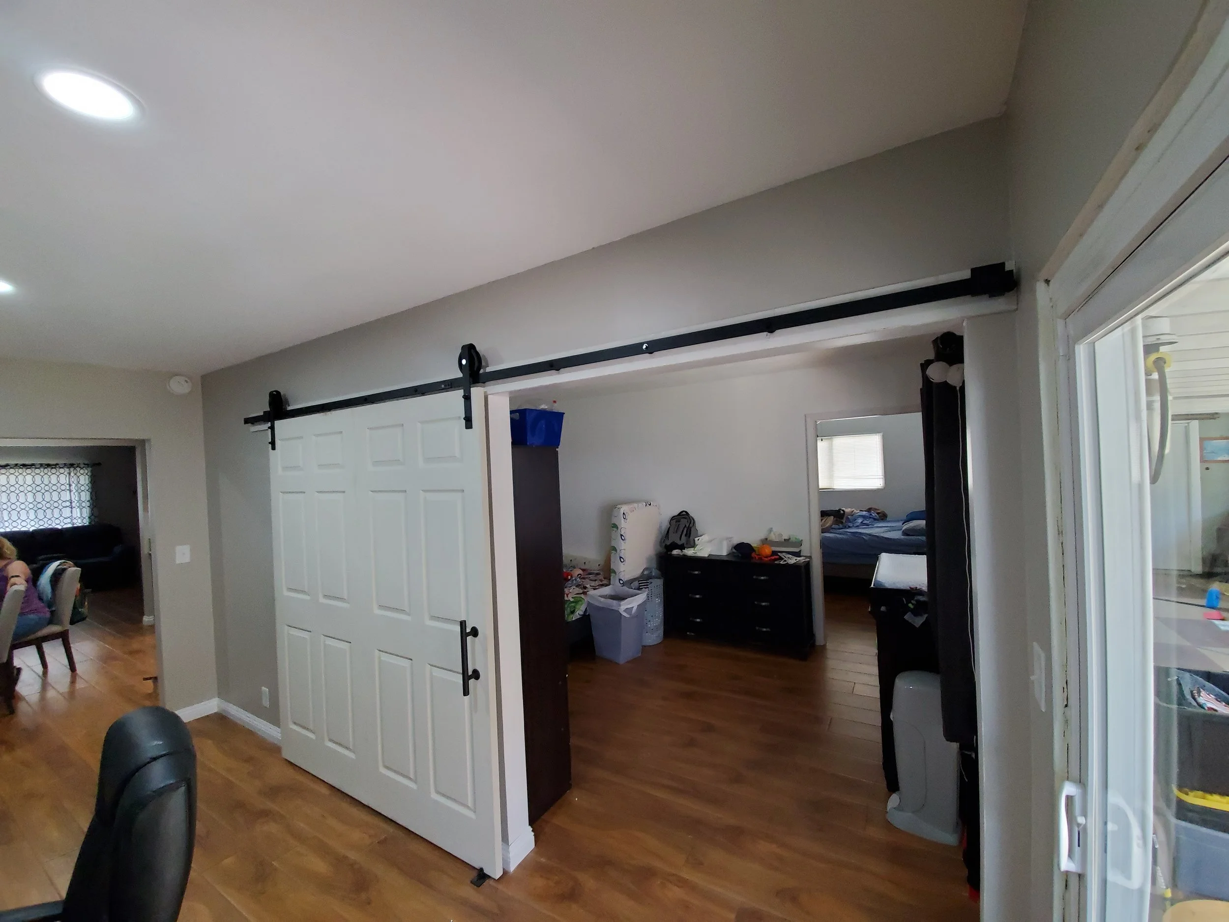 Interior view of a room with wooden flooring, a white sliding barn door on a black track, a black chair, and a partial view of another room with a bed and various items.