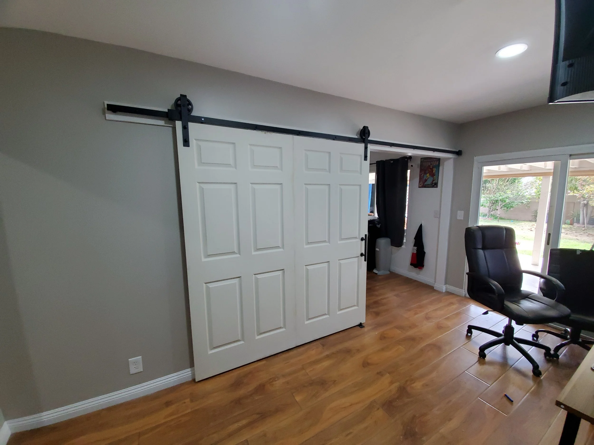 A room with light gray walls, a sliding barn door painted white with black hardware, a window with black curtains, and a patio door leading outside. There's a black office chair and a small poster on the wall.