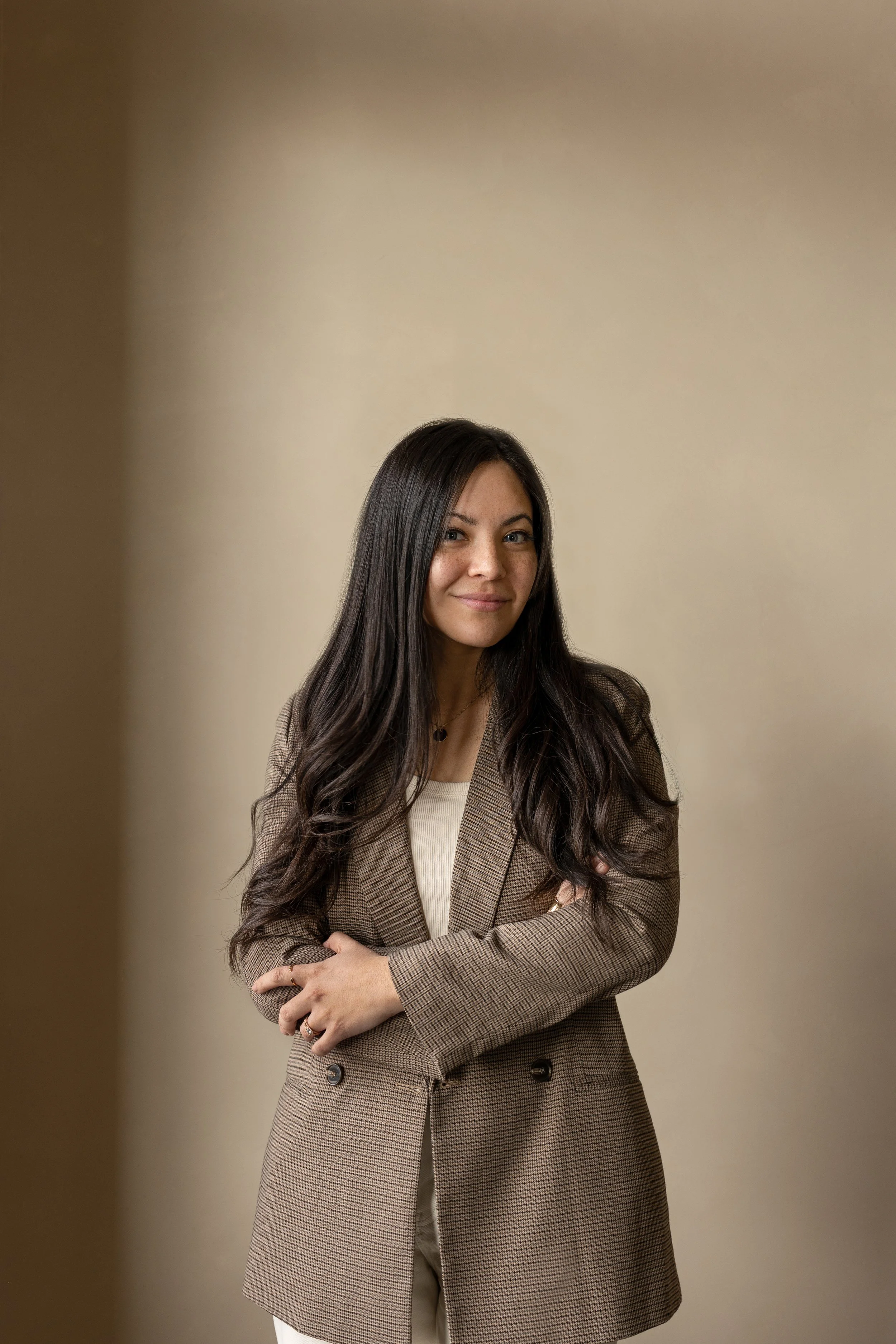 Portrait of Pam Miyamoto smiling, wearing a plaid brown blazer against a soft neutral background.