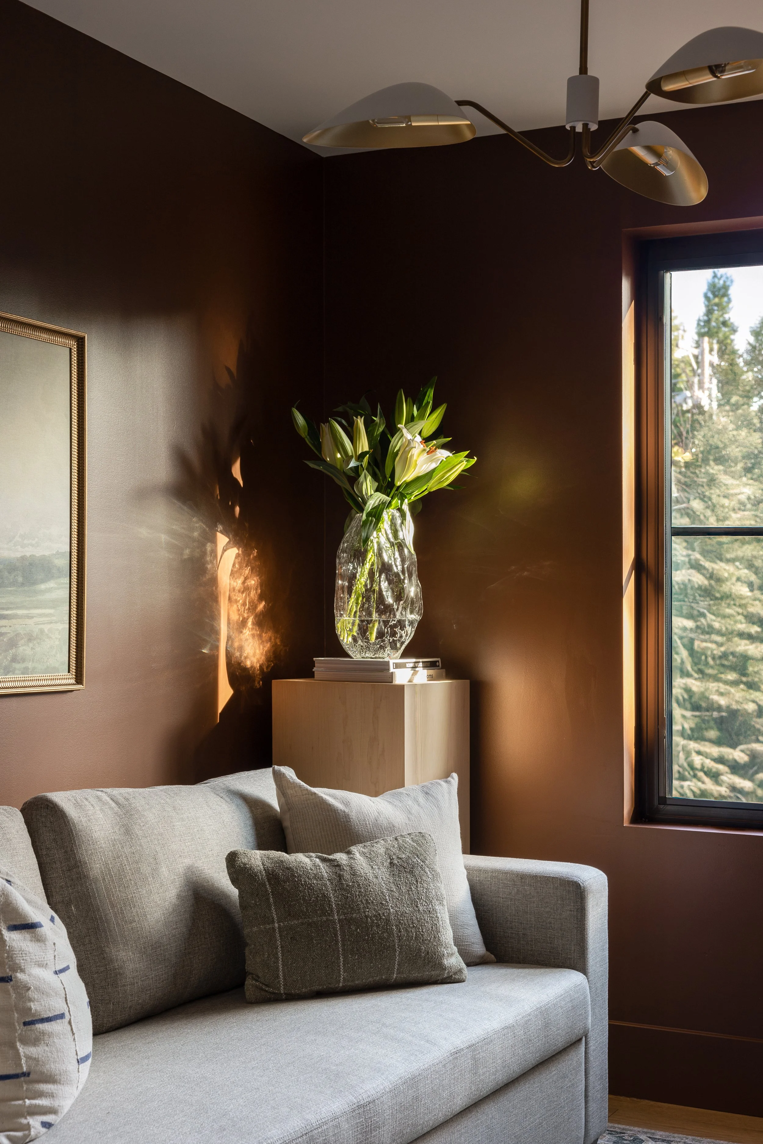 Dark brown moody office with art above sofa, glass vase in corner, brown walls, and modern pendant