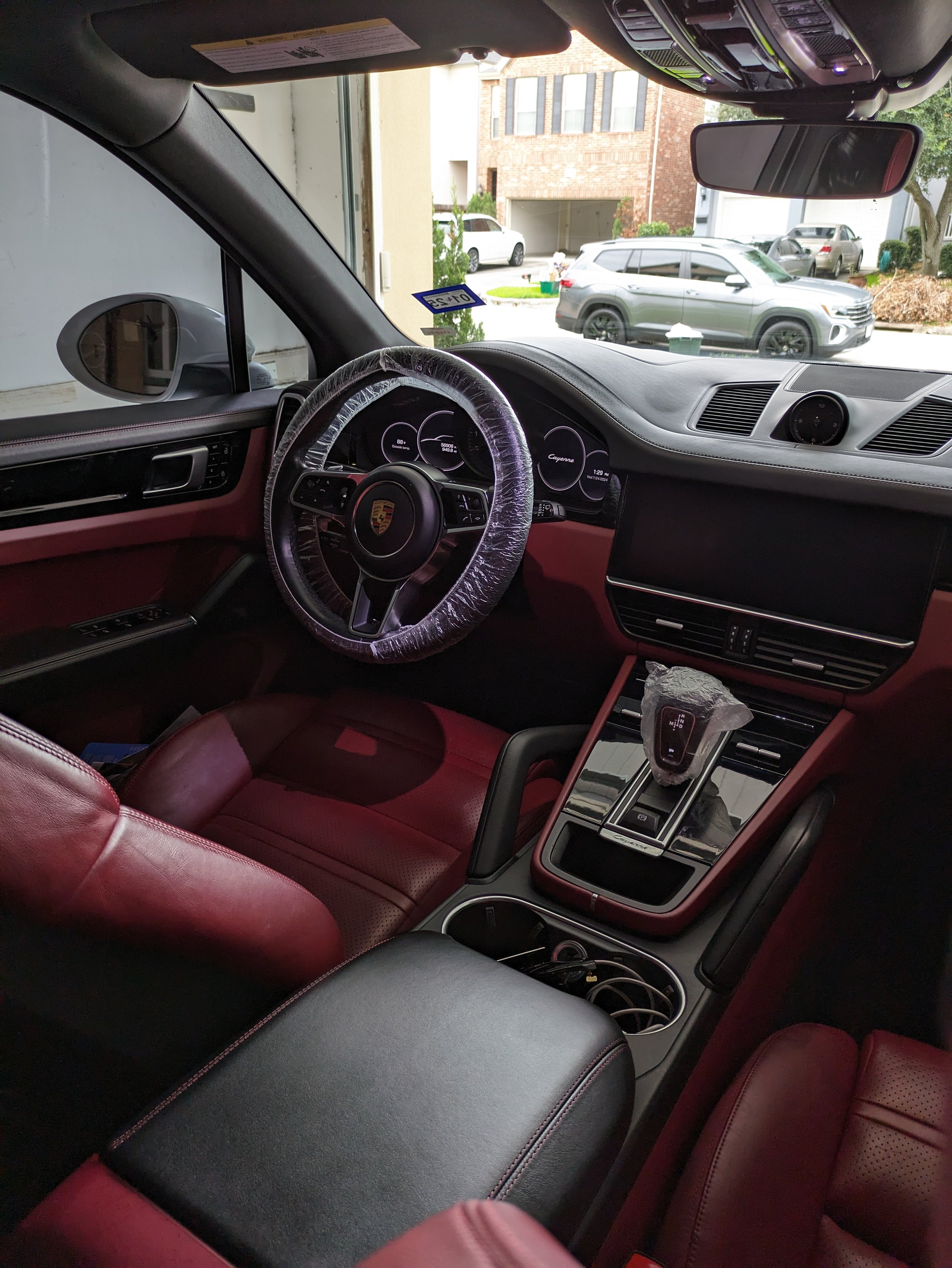 Interior of a luxury car with red leather seats, a wrapped steering wheel, and a central console featuring a gear shift. The dashboard displays several dials and a digital screen. The outside view shows a residential street with parked cars.