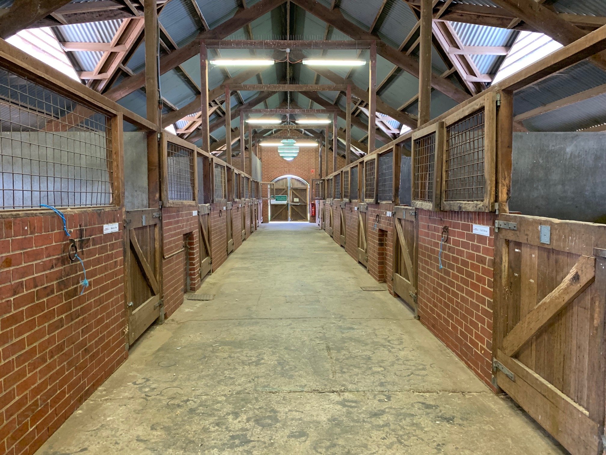 Inside of a barn with a central aisle flanked by stalls on both sides, brick walls, wooden partitions, and a gabled metal roof.