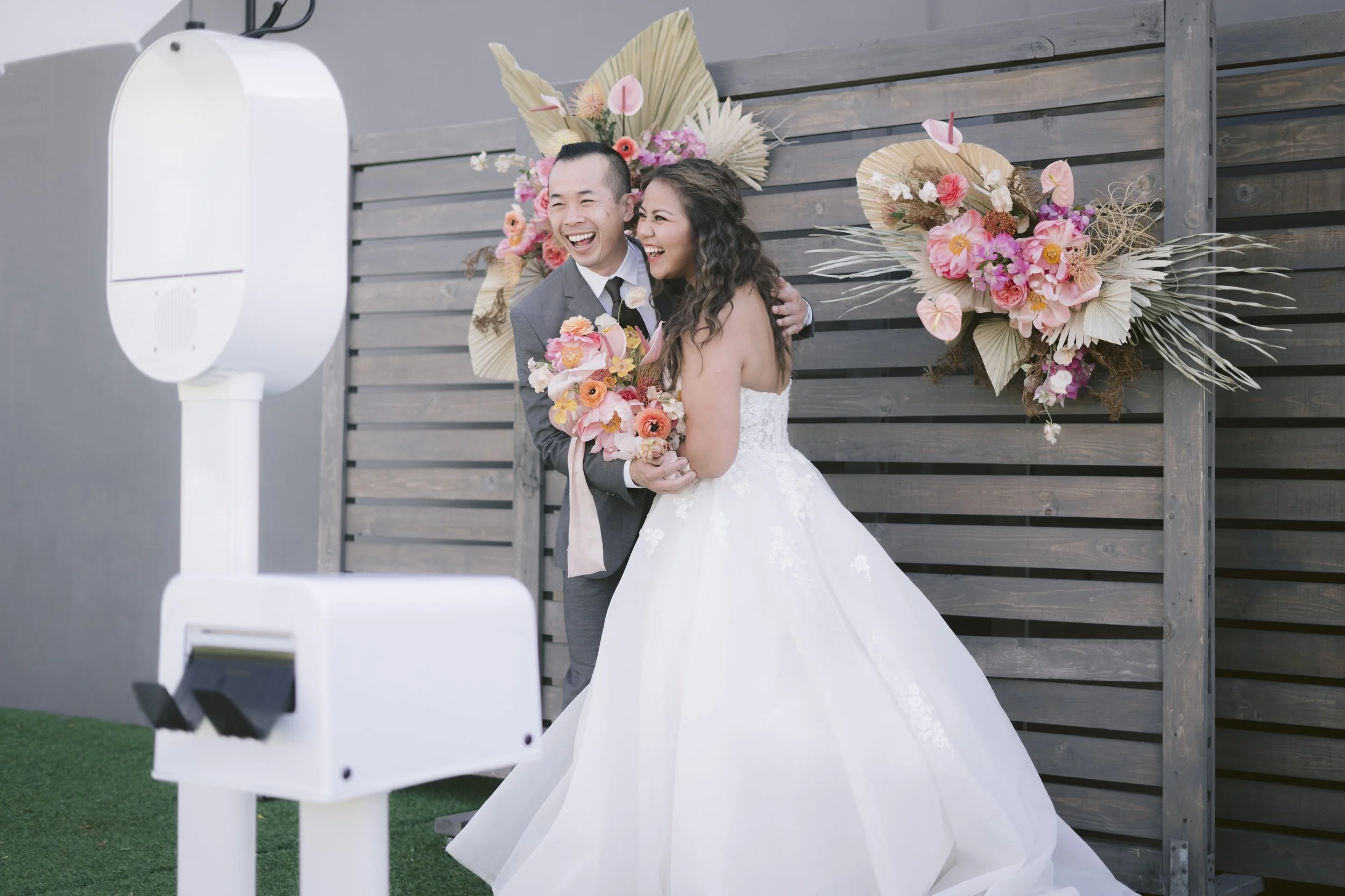 A bride and groom smiling and posing in front of a wooden fence with floral decorations, standing next to a modern photo booth.