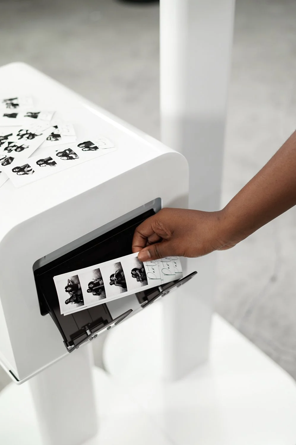 Person removing photo strip from printer, with multiple black and white photo strips on top of a white device.