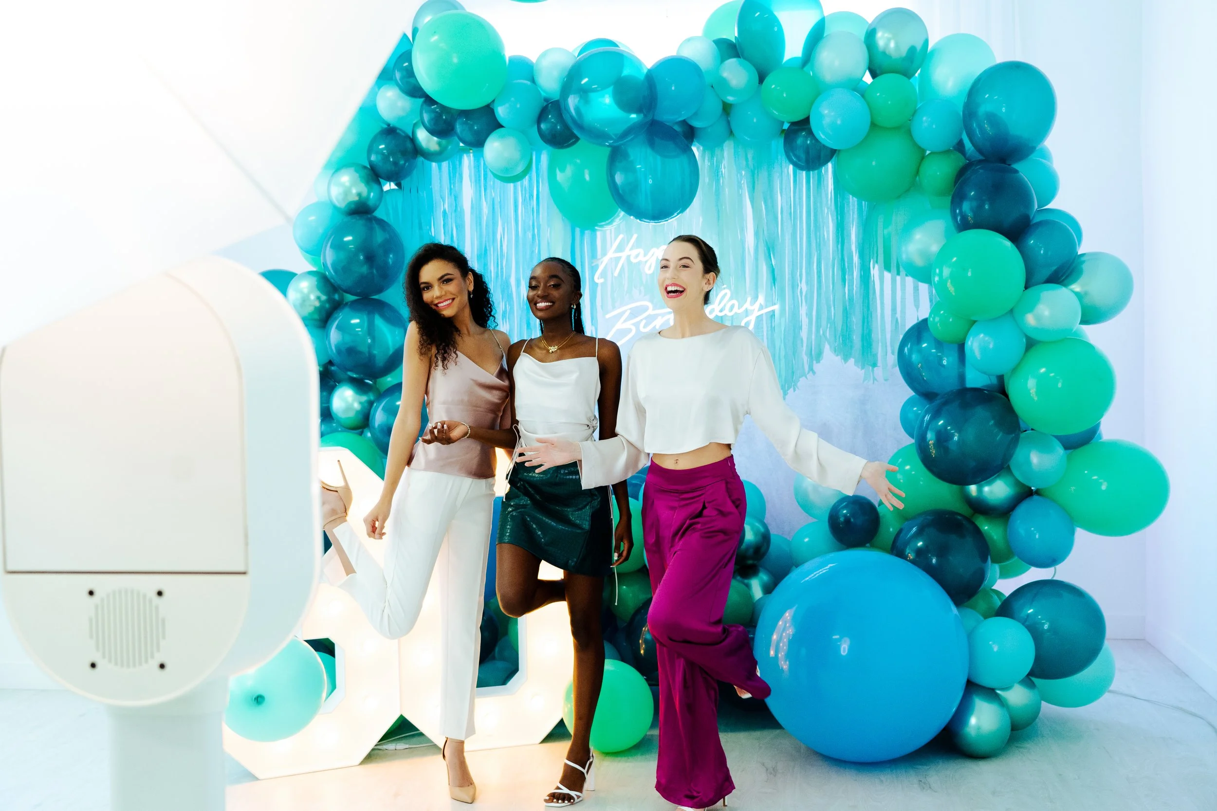 Three women pose joyfully at a birthday party in front of a teal and blue balloon arch, with "Happy Birthday" neon sign in the background.