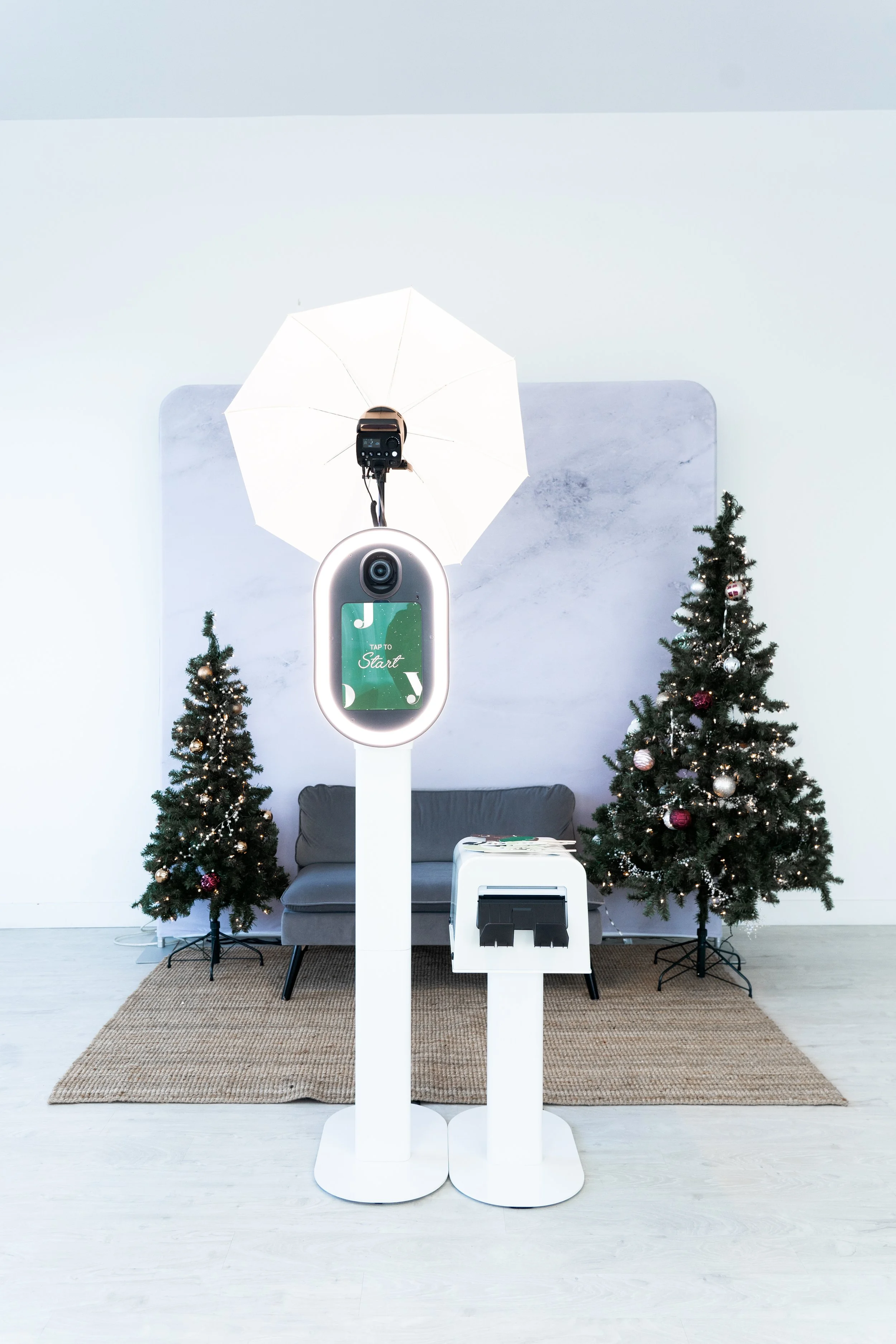 Photography setup with a photo booth, two Christmas trees, a backdrop, and a sofa. The photo booth has a ring light, a camera, and a "Tap to Start" sign. A printer is placed beside it.