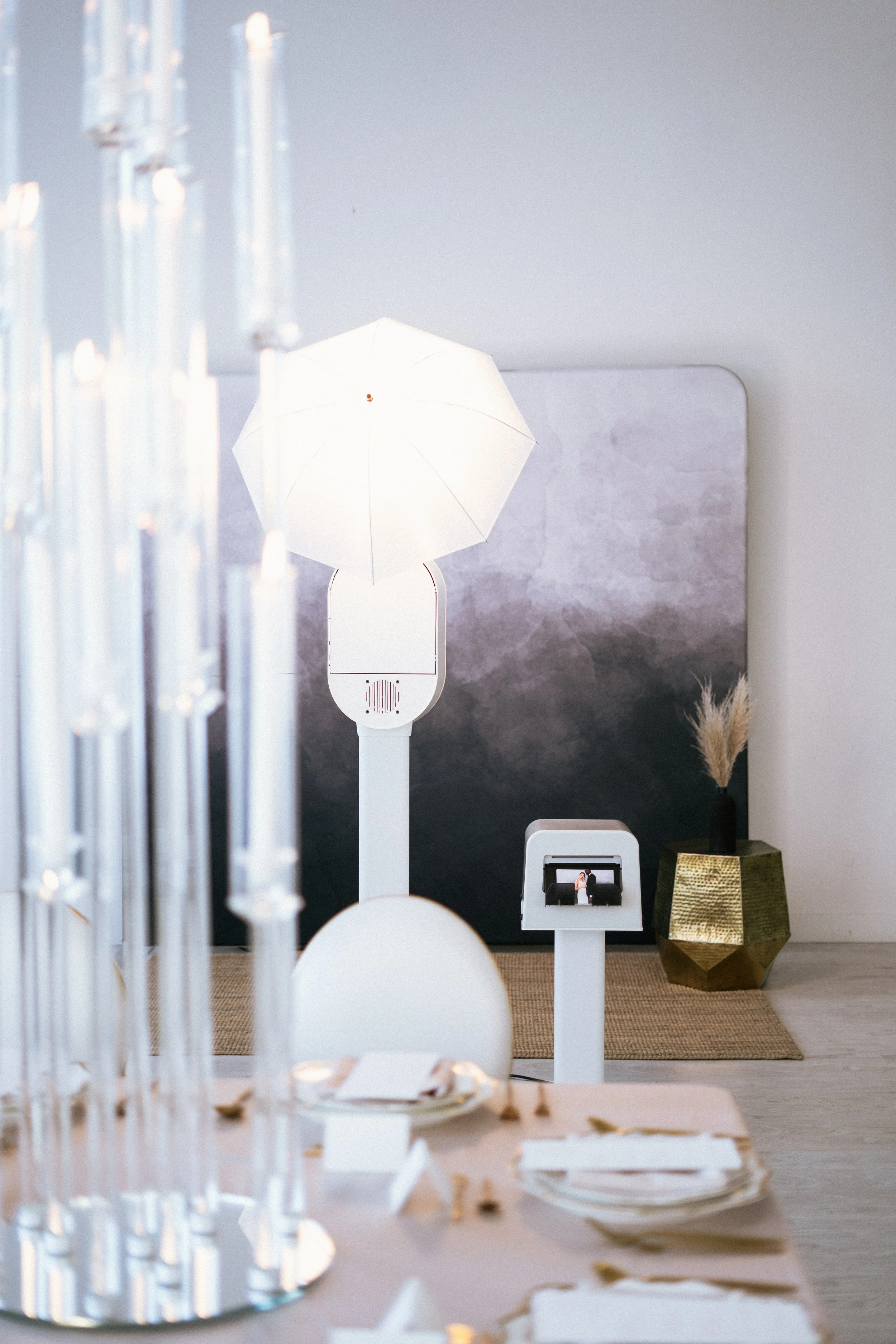 Elegant table setting with glass candleholders, a photo booth with umbrella light, and decorative elements in a modern venue.