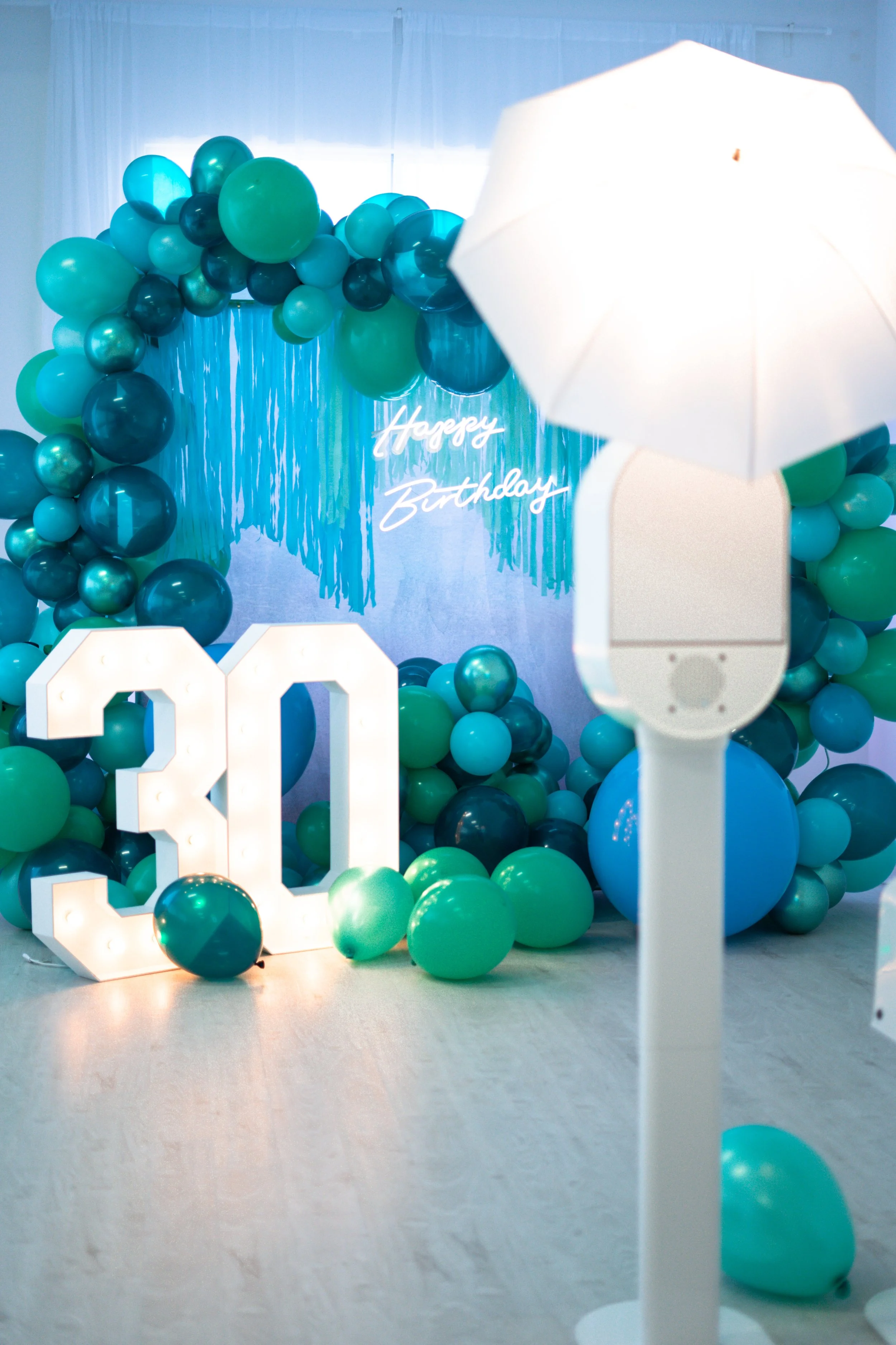 Birthday party decoration with blue and green balloon arch, light-up '30' sign, and 'Happy Birthday' neon sign.