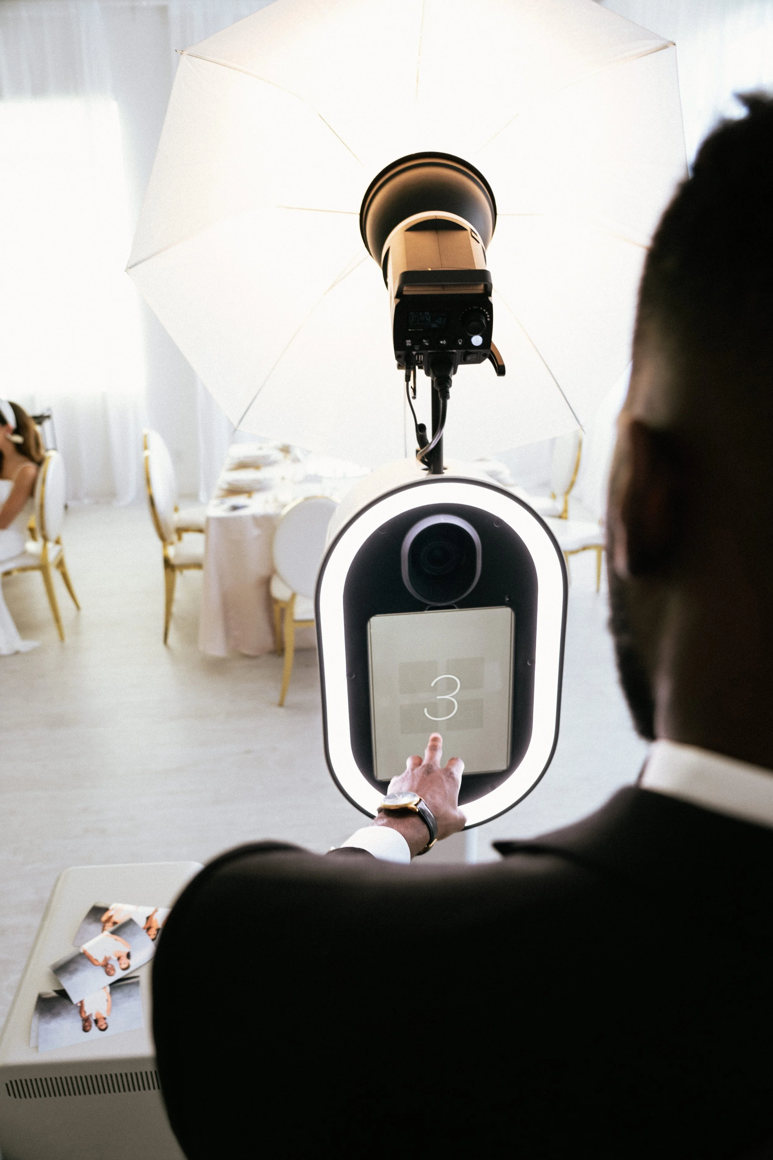Person using a photo booth touchscreen at an event with a camera and light setup, pressing a button displaying the number 3, surrounded by elegant seating and decor.