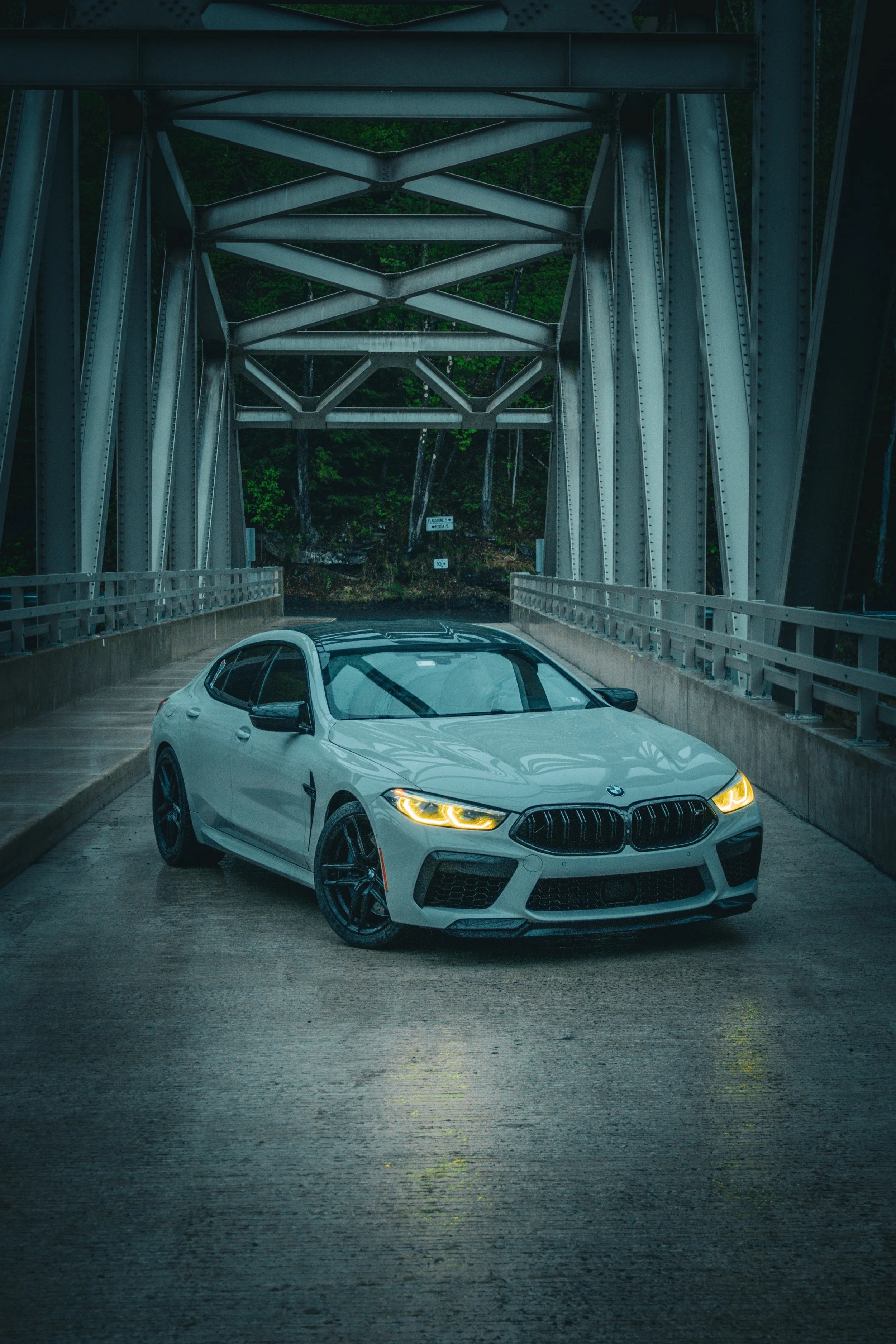A gray BMW car with black rims driving on a bridge with steel trusses and green trees in the background.