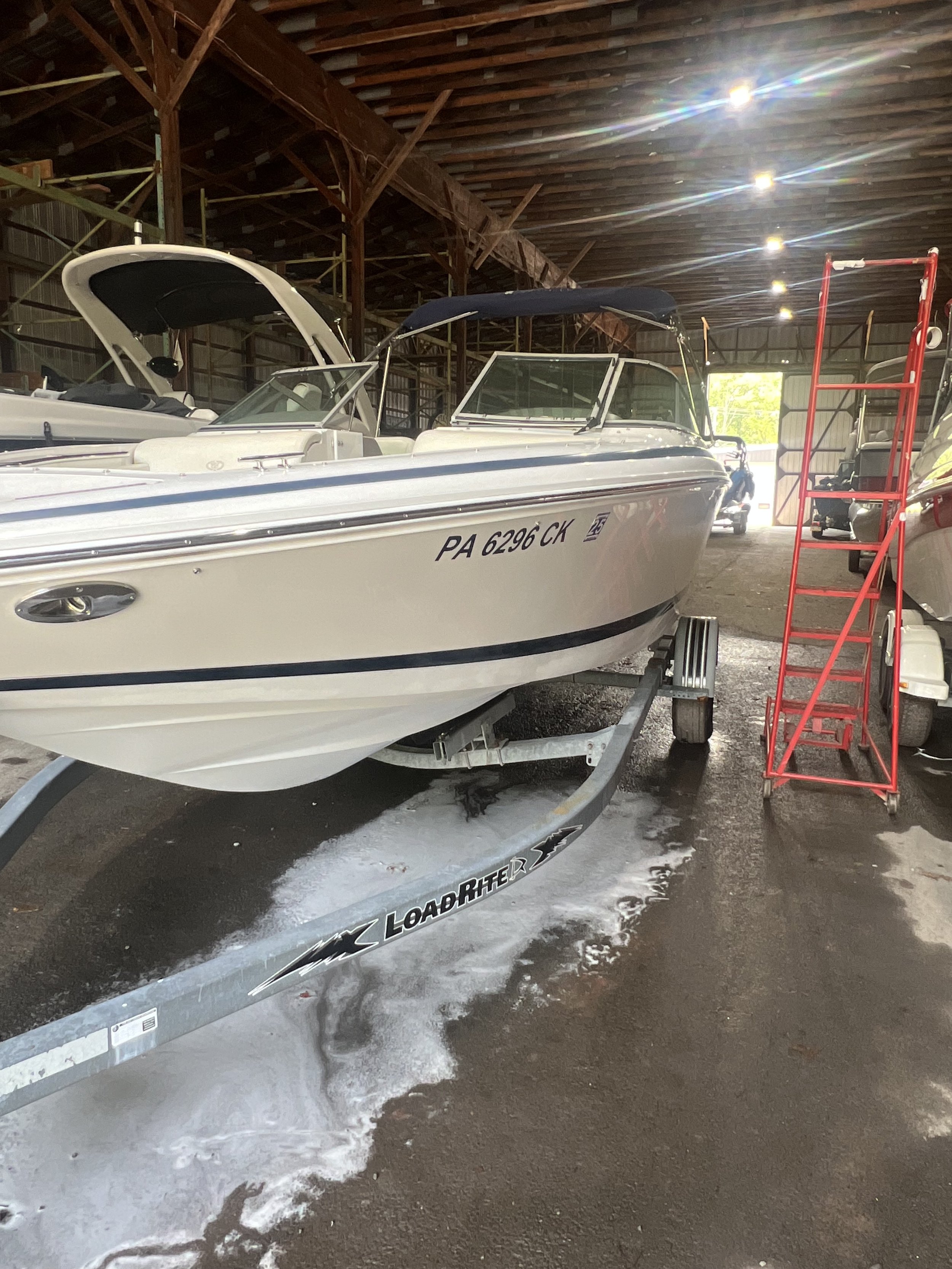A white speedboat on a trailer inside a boat storage or maintenance facility, with foam on the concrete floor and a red ladder beside it.