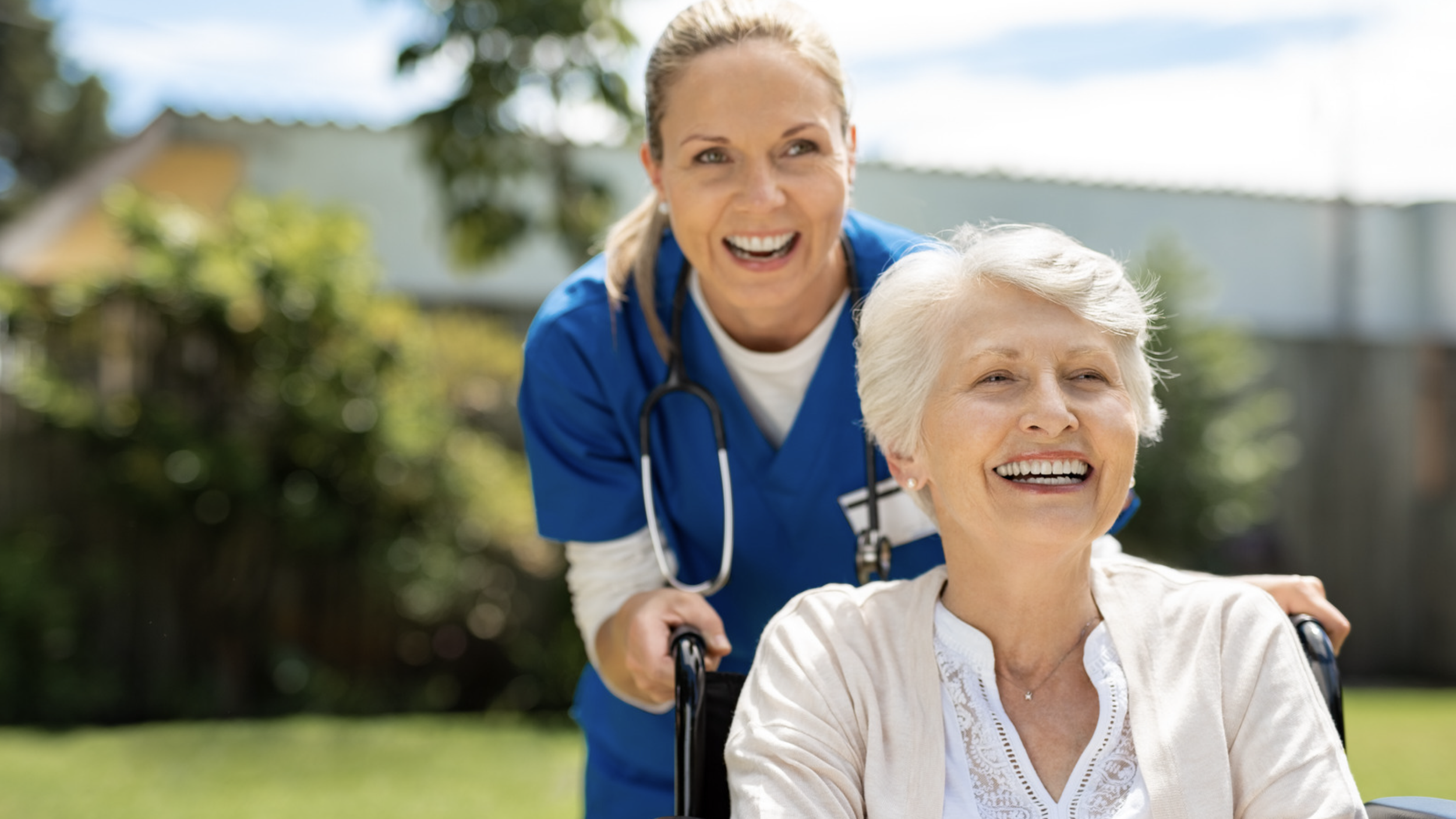 A healthcare worker pushing an elderly woman in a wheelchair outdoors on a sunny day, both smiling.