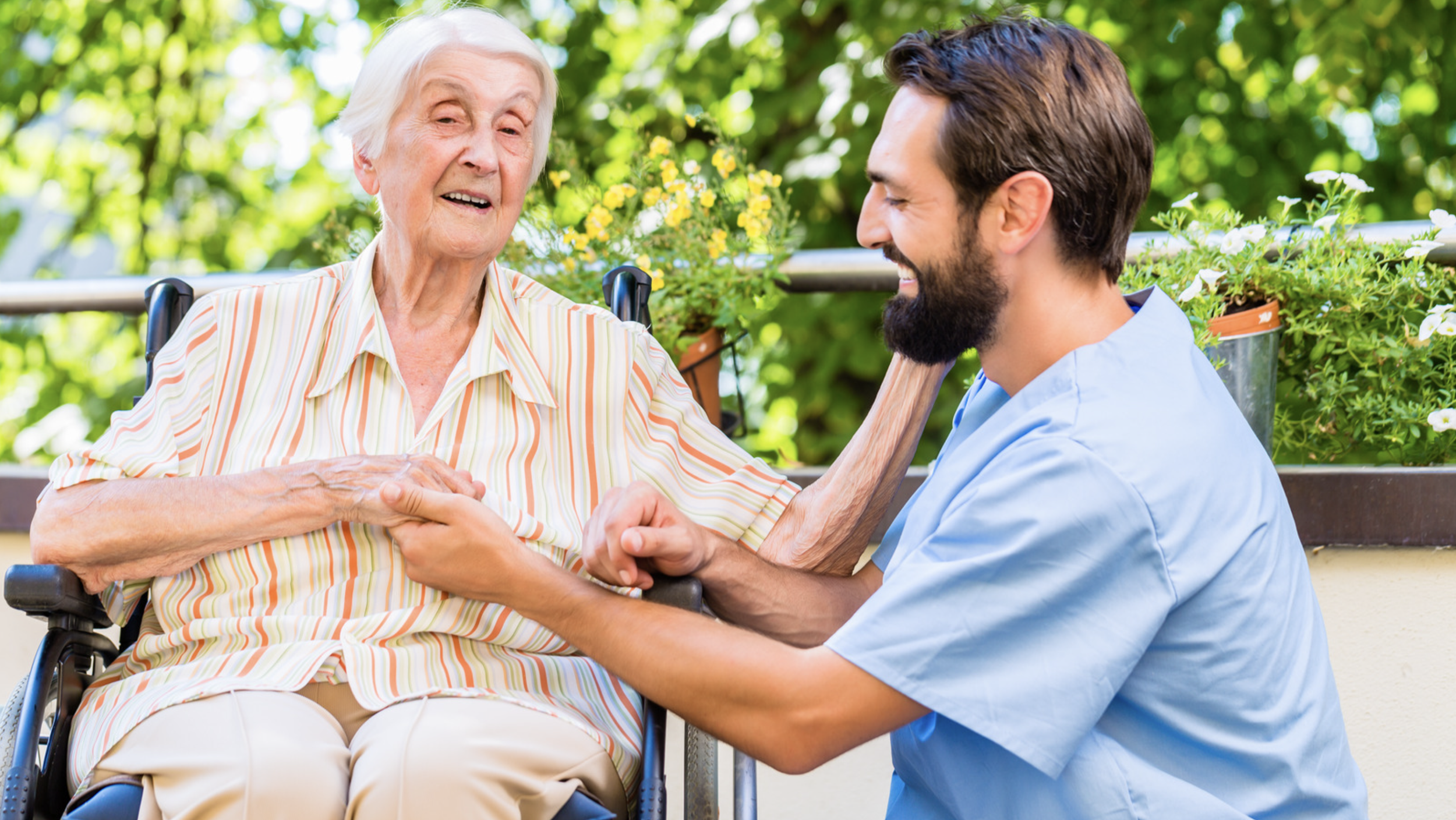A young male caregiver with a beard, wearing light blue scrubs, kneeling and smiling while holding hands and talking to an elderly woman in a wheelchair, outdoors with green foliage and flowers.