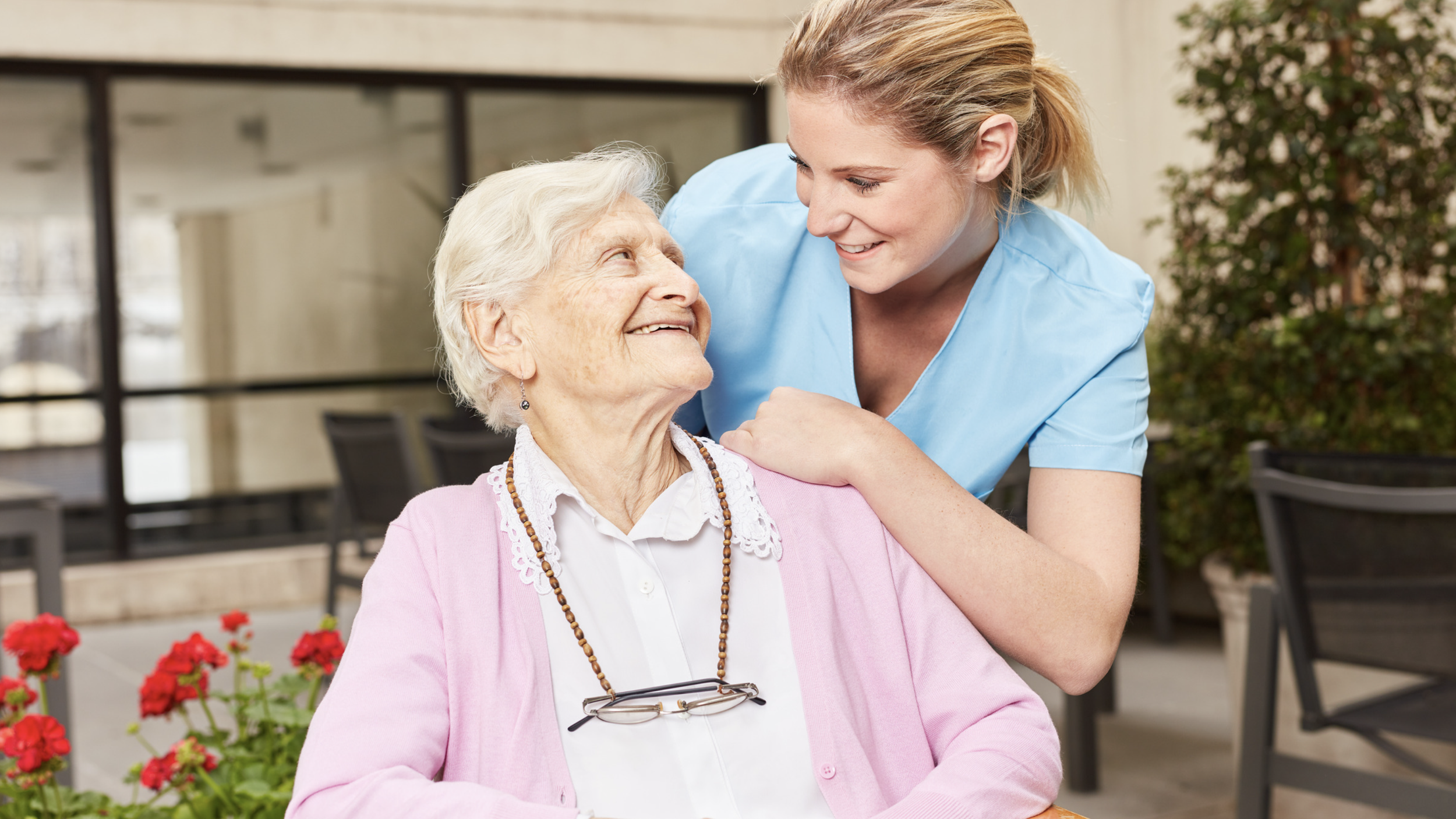 A young female caregiver in light blue uniform smiling at an elderly woman with white hair, wearing a pink cardigan and glasses, on a terrace with potted plants and flowers.
