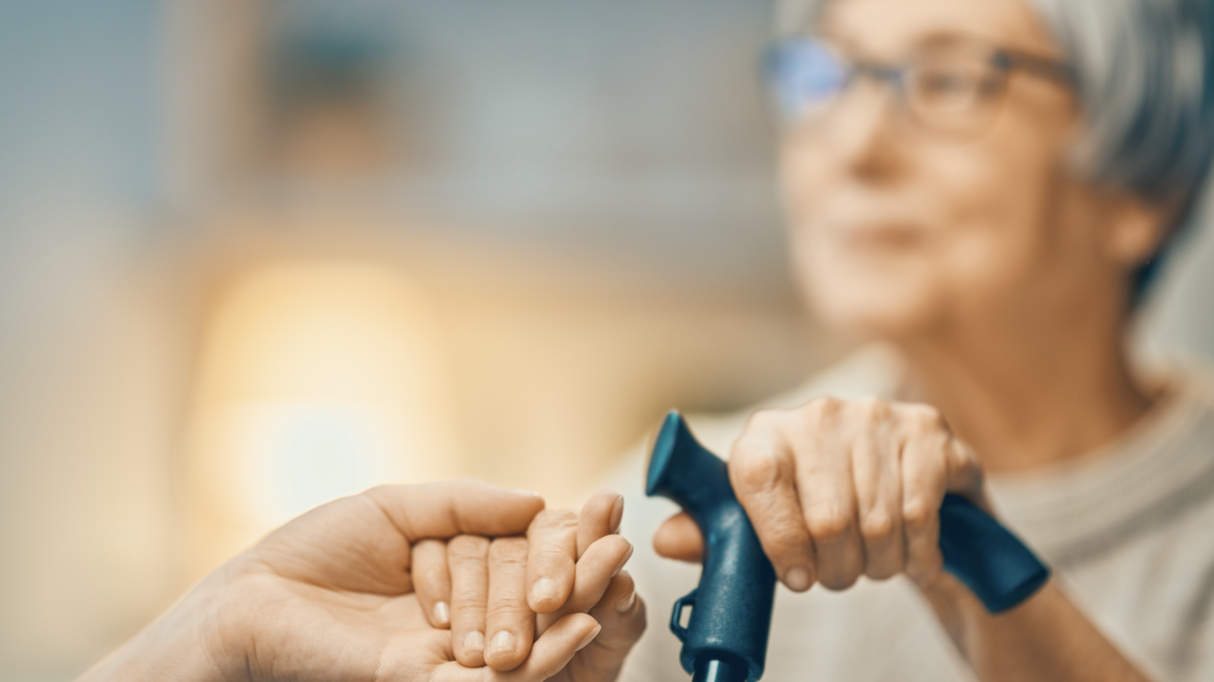 An elderly woman with glasses and headphones holding a black cane, being assisted by a person with their hand gently holding her hand.
