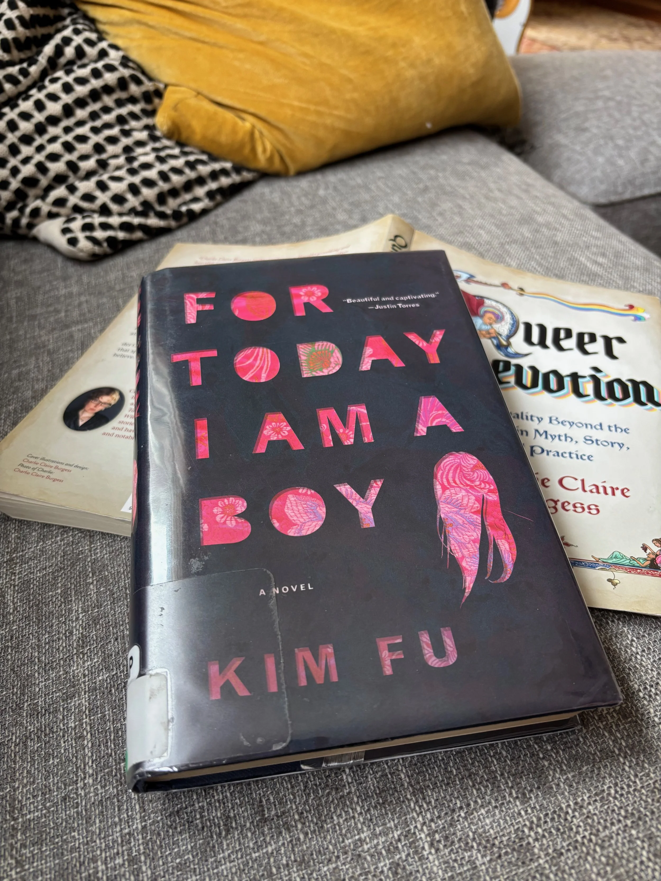 A hardover library copy of For Today I Am a Boy by Kim Fu lays on top of on open, face down copy of Queer Devotion by Charlie Claire Burgess on the blogger's gray sofa