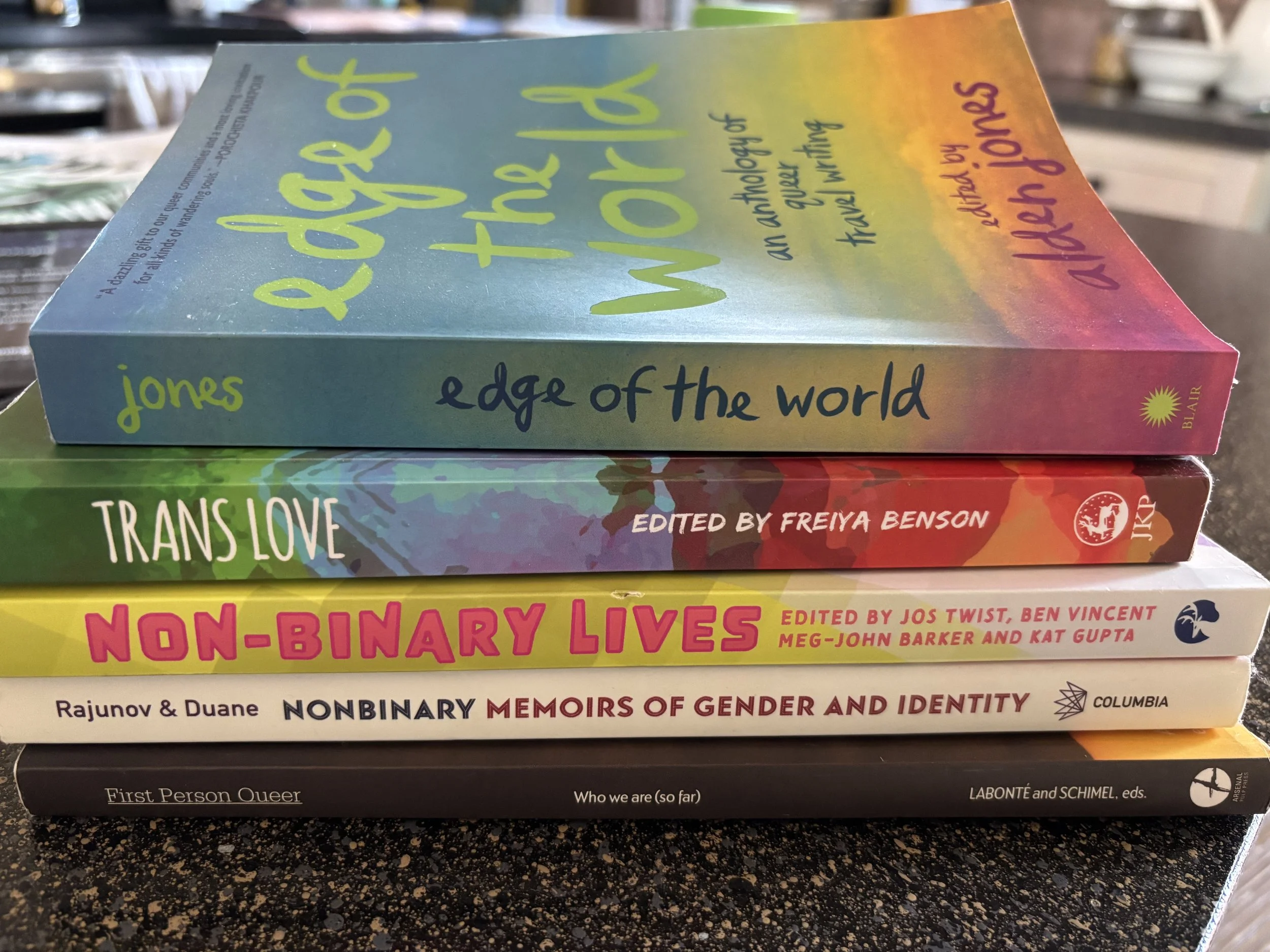 a rainbow-colored paperback book sits on top of four other paperback essay anthologies