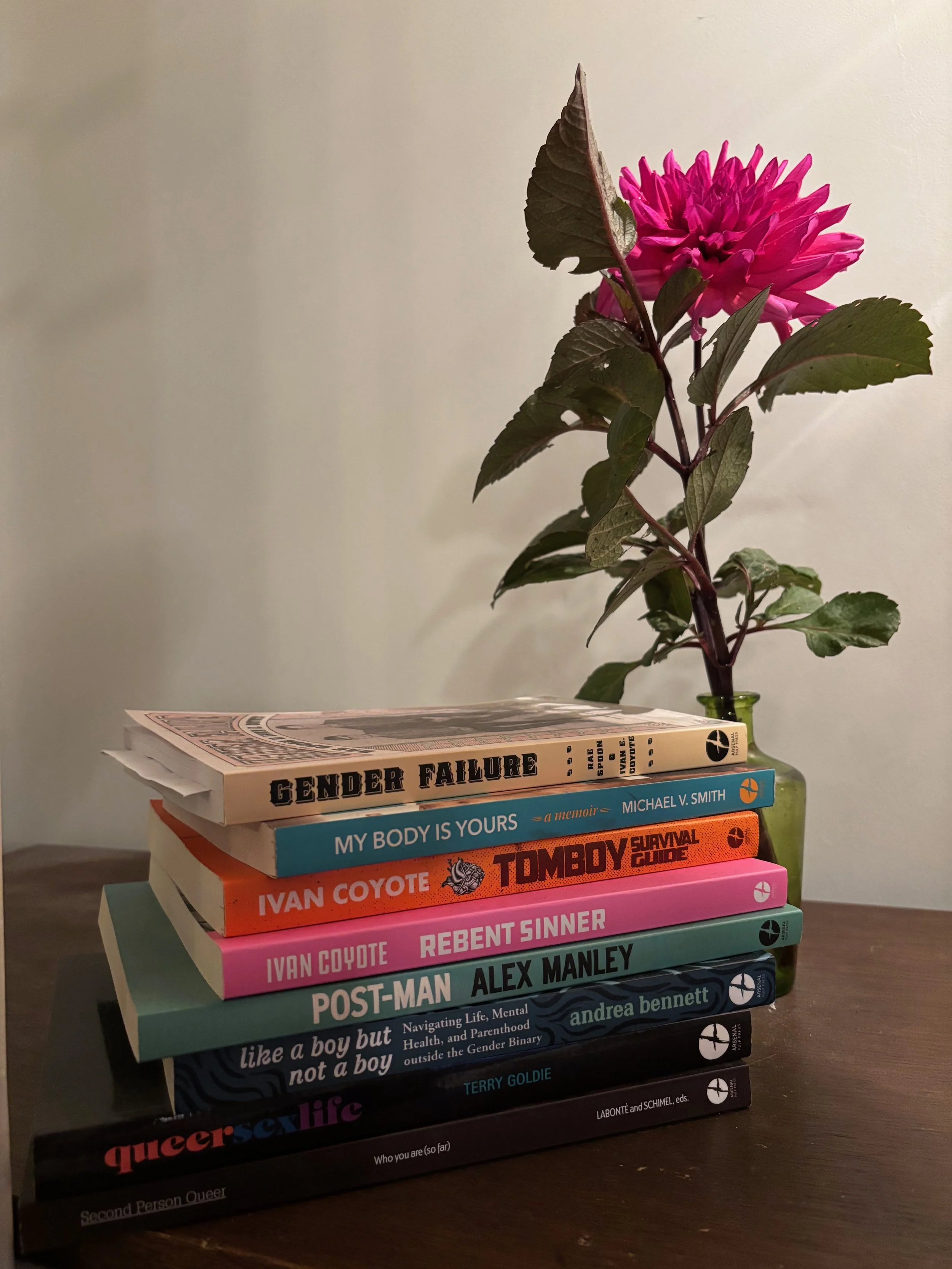 a stack of  eight queer paperback books published by arsenal pulp press sits in front of a fuschia colored late season cut dahlia in a small green bottle